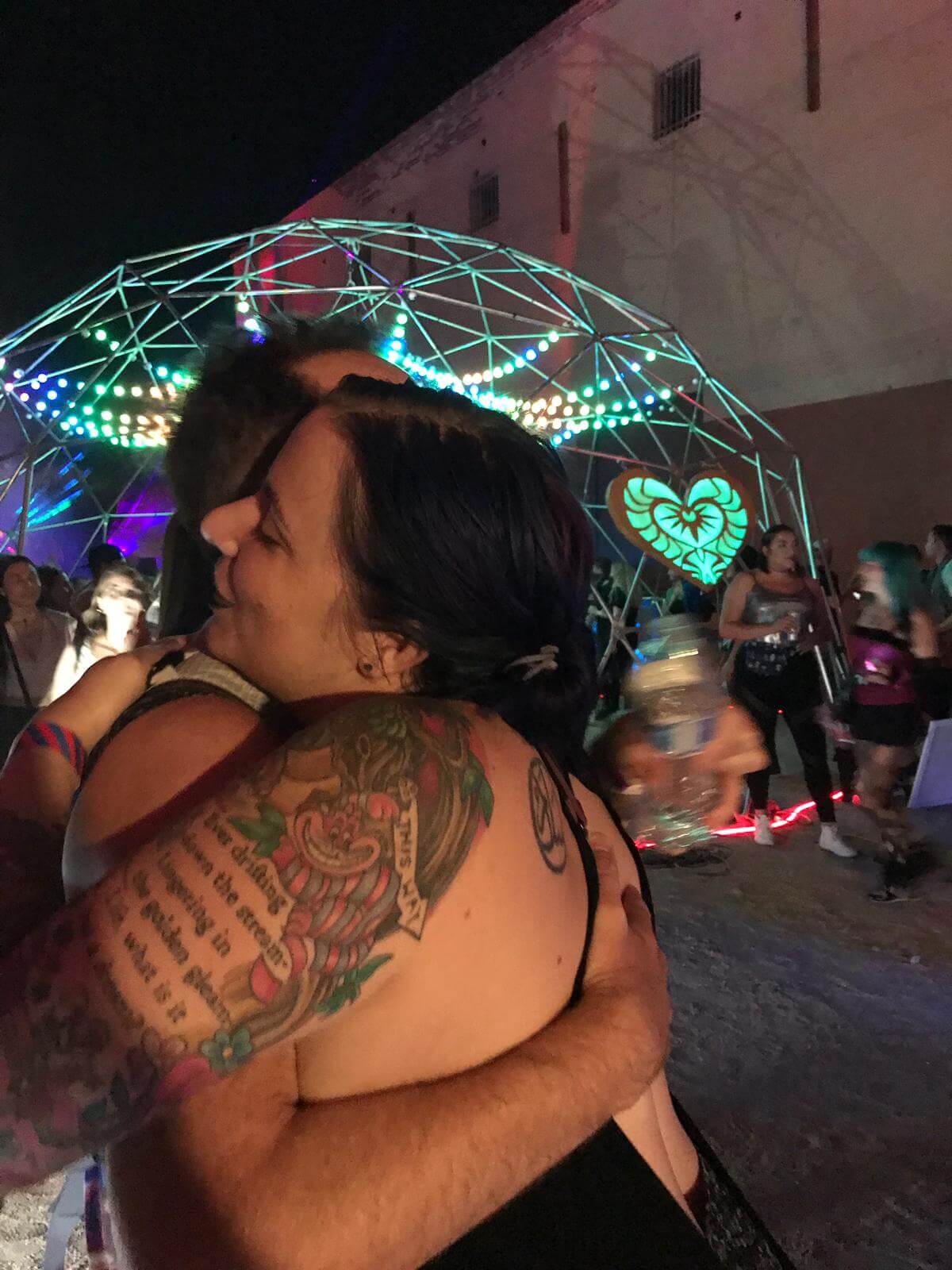 Two people embrace at a nighttime outdoor event with colorful, geometric LED lights in the background. One person has visible tattoos on their arm, and other attendees can be seen enjoying the festive atmosphere.