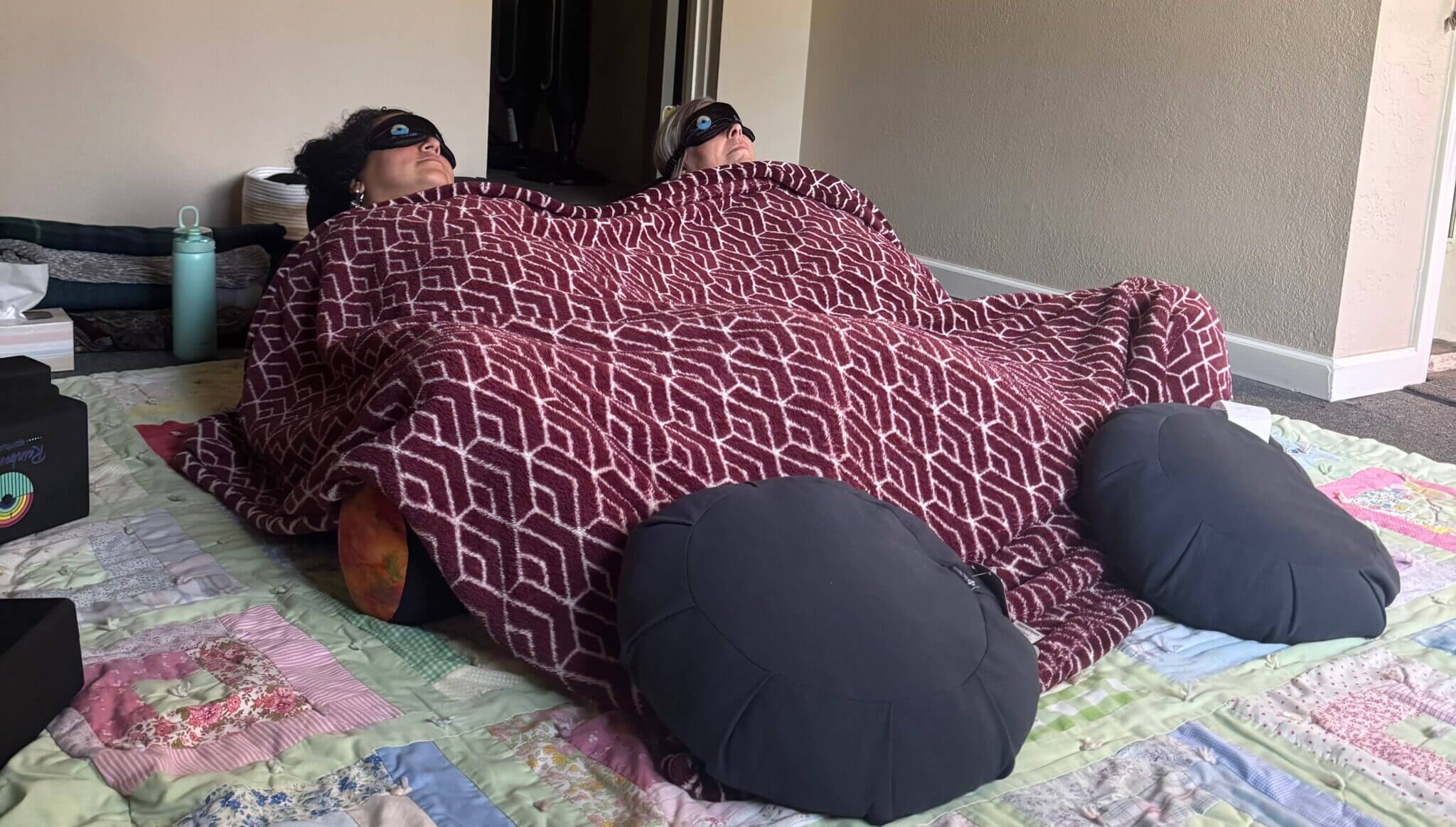 Two people lie side by side on a quilted blanket, covered with a red patterned blanket up to their chests. Both wear black eye masks, with their feet propped up on large black round cushions.