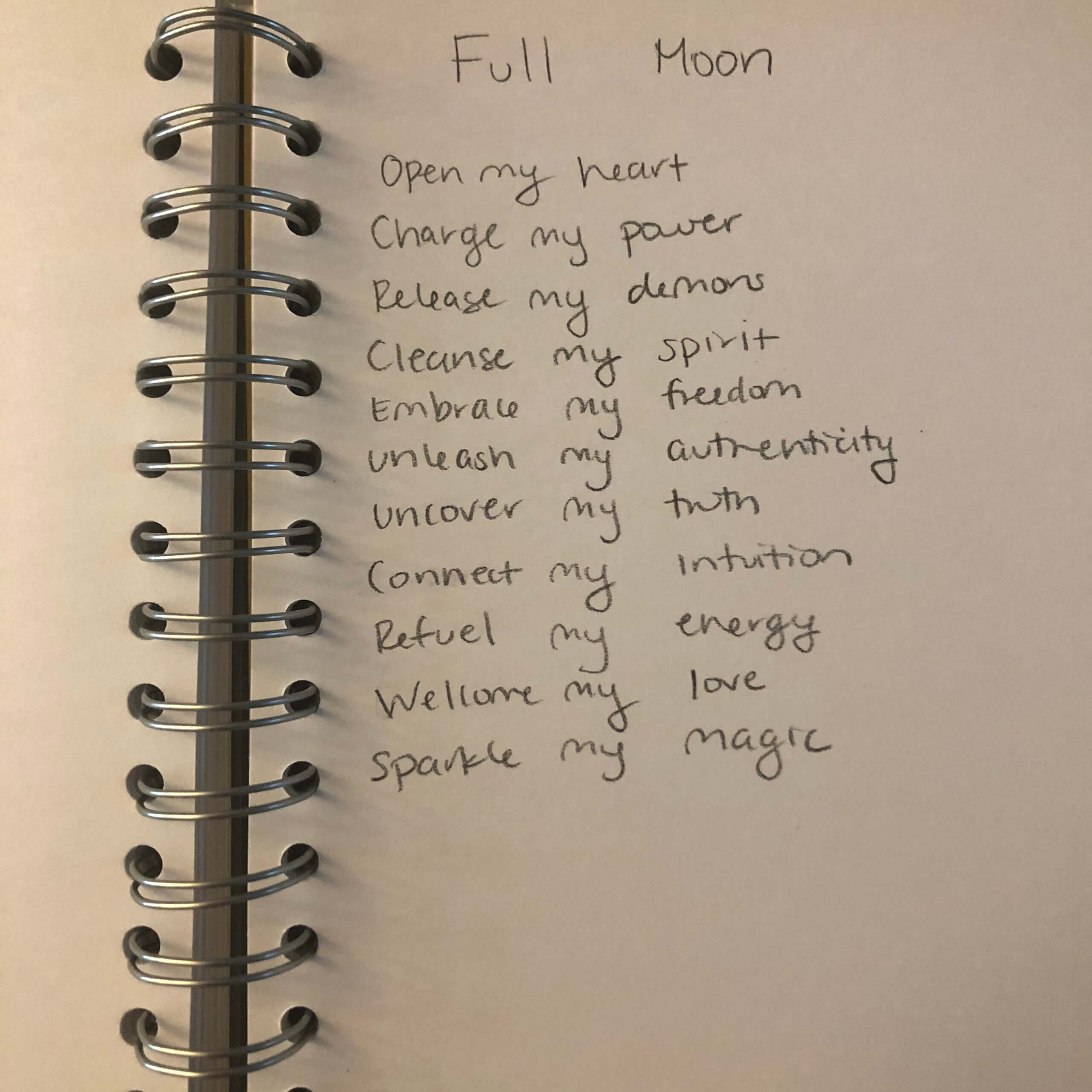 A handwritten poem titled Full Moon on a spiral notebook page, listing affirmations such as Open my heart, Charge my power, Release my demons, and others in neat, cursive writing.