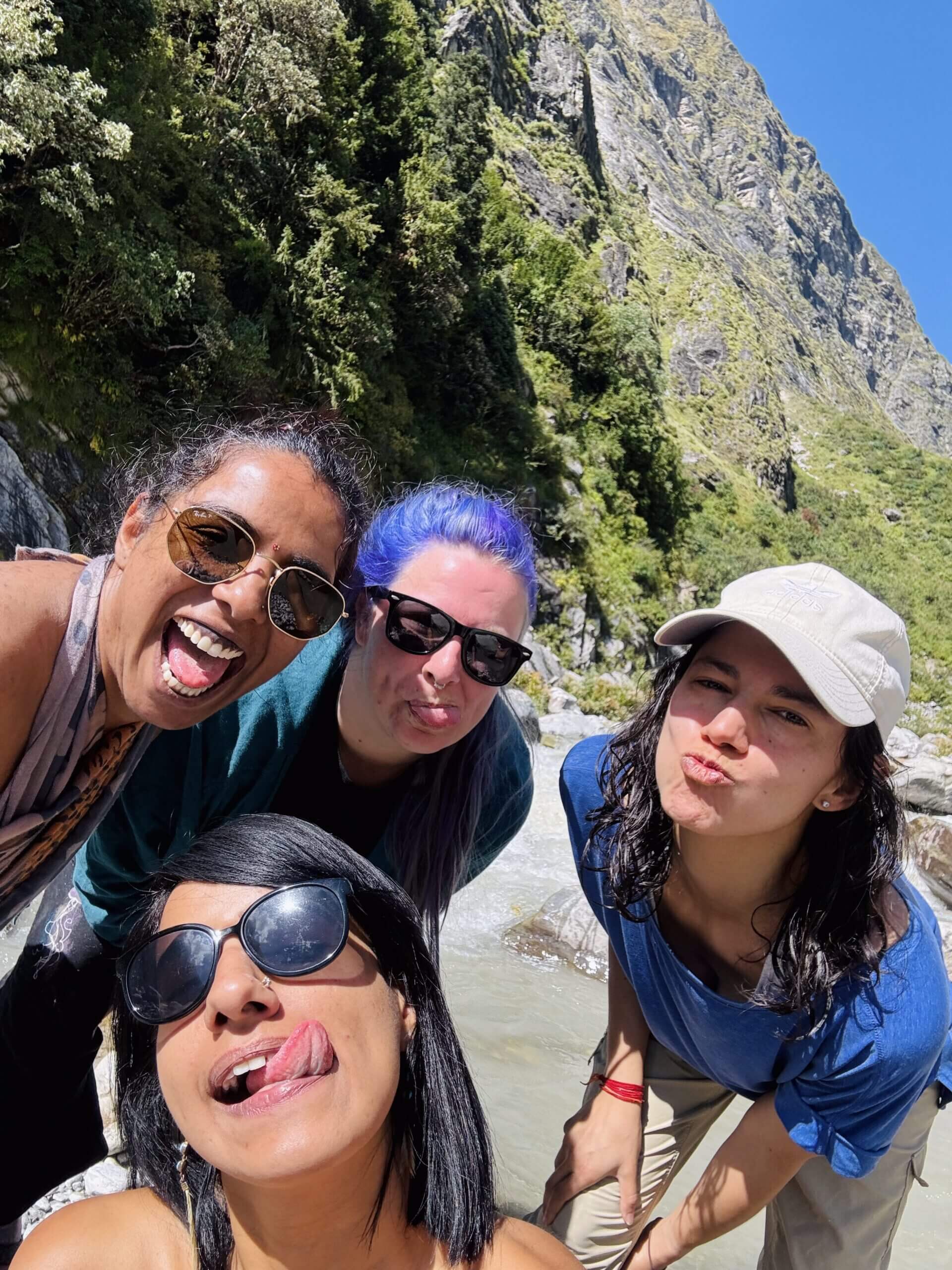 Four people outdoors at the base of a mountain, all smiling and making playful faces at the camera. They wear sunglasses and casual clothes, with greenery and a rocky hill in the background under a clear blue sky.