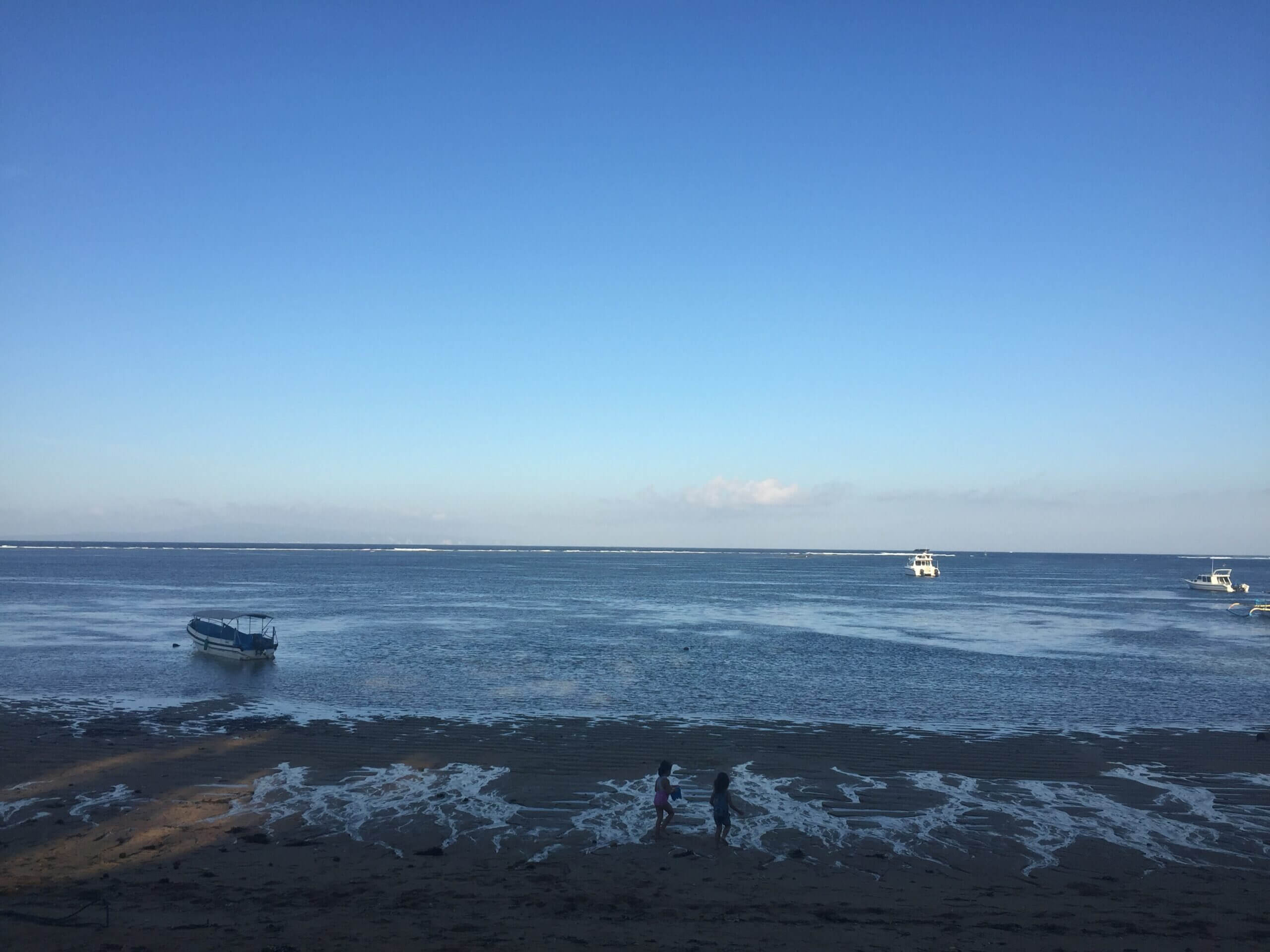 A tranquil beach scene with two people walking on the sand near gentle waves. Three small boats float on the calm blue sea under a clear sky.