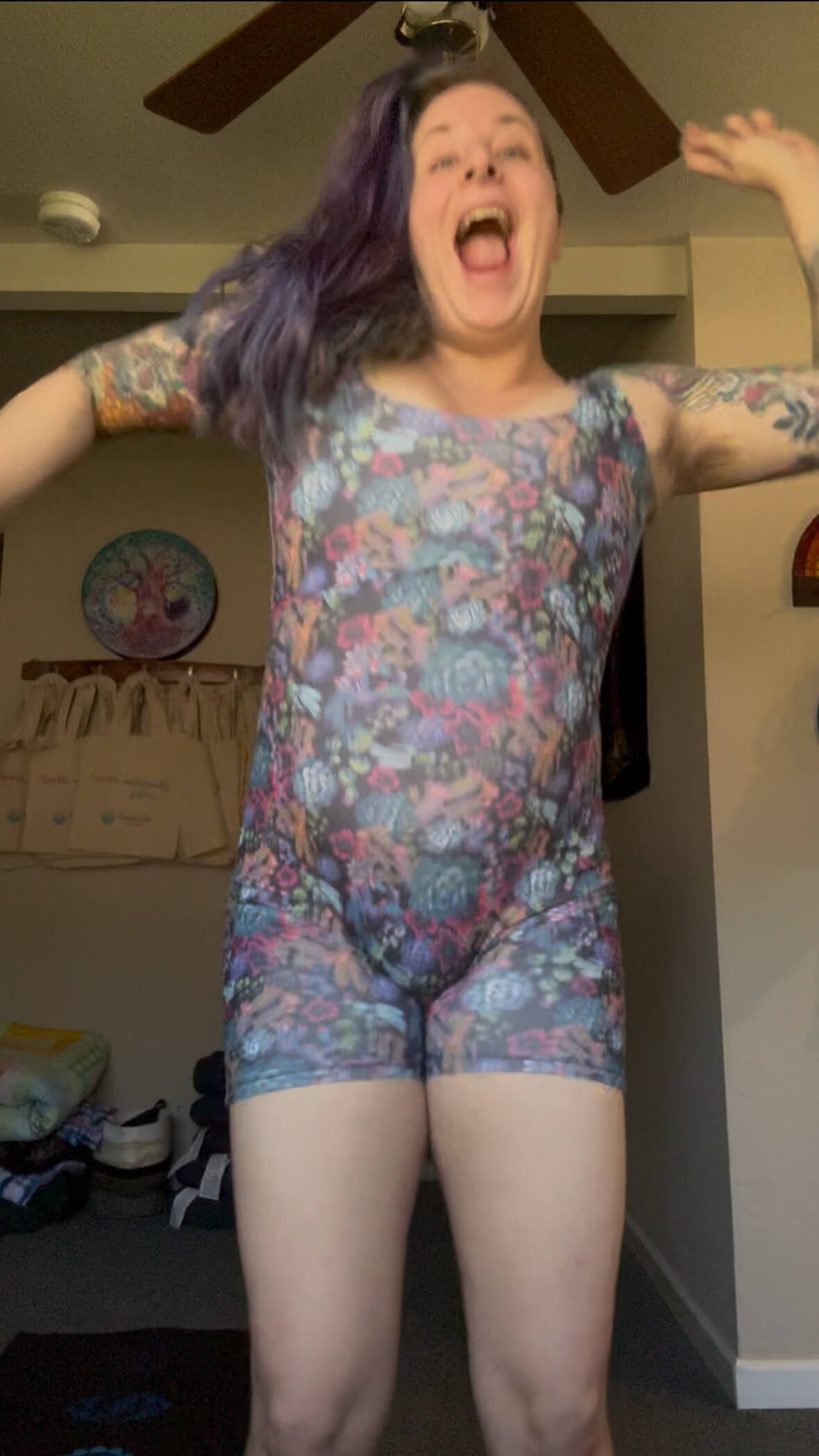 A person with long purple hair and tattoos is joyfully jumping indoors, wearing a colorful floral romper. The background shows a decorated wall, some bags, and household items.