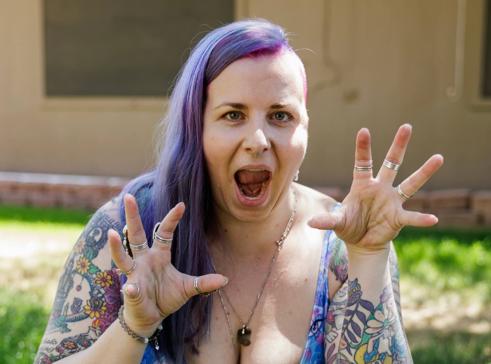 A person with purple hair and tattoos, wearing rings, poses outdoors with their hands raised like claws and mouth open in a playful expression.