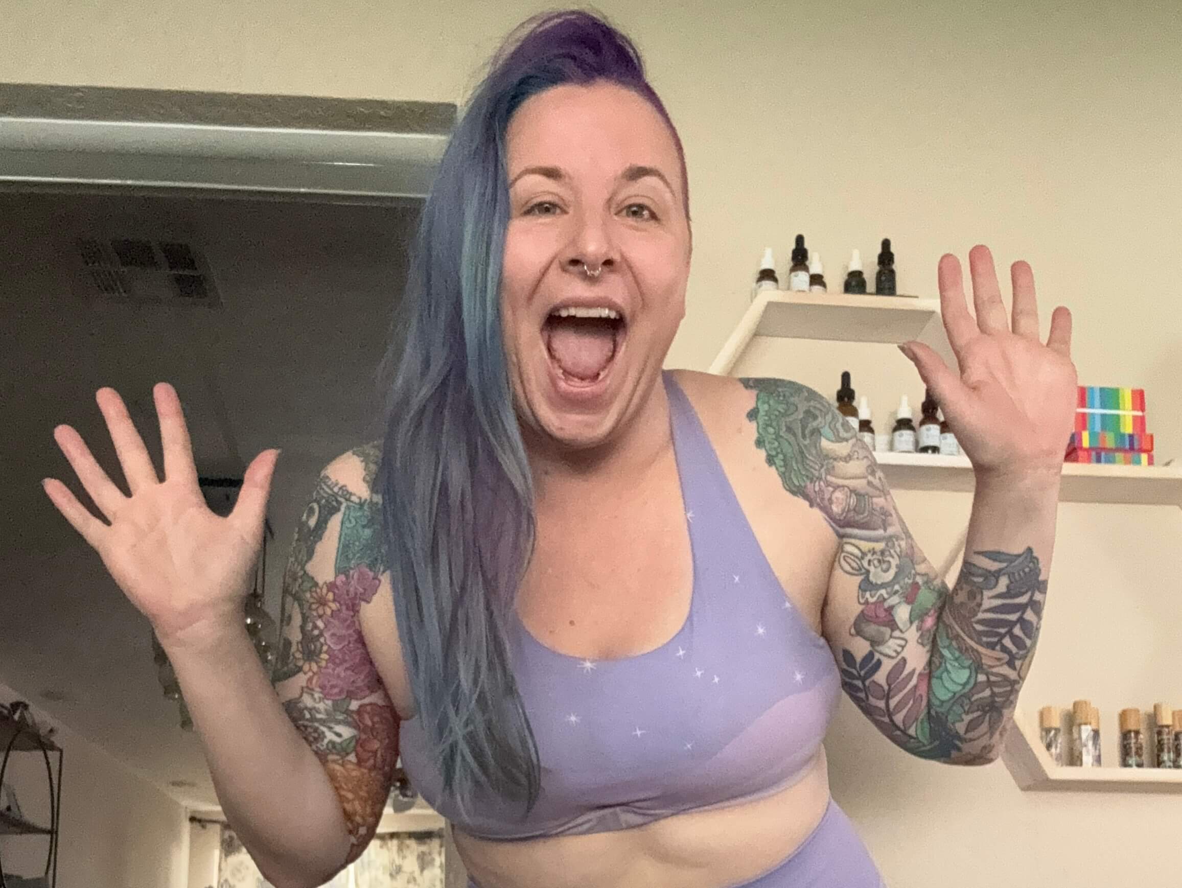 A person with long, purple-and-blue hair and colorful tattoos smiles widely with hands up, wearing a lavender sports bra, standing indoors with shelves and bottles visible in the background.