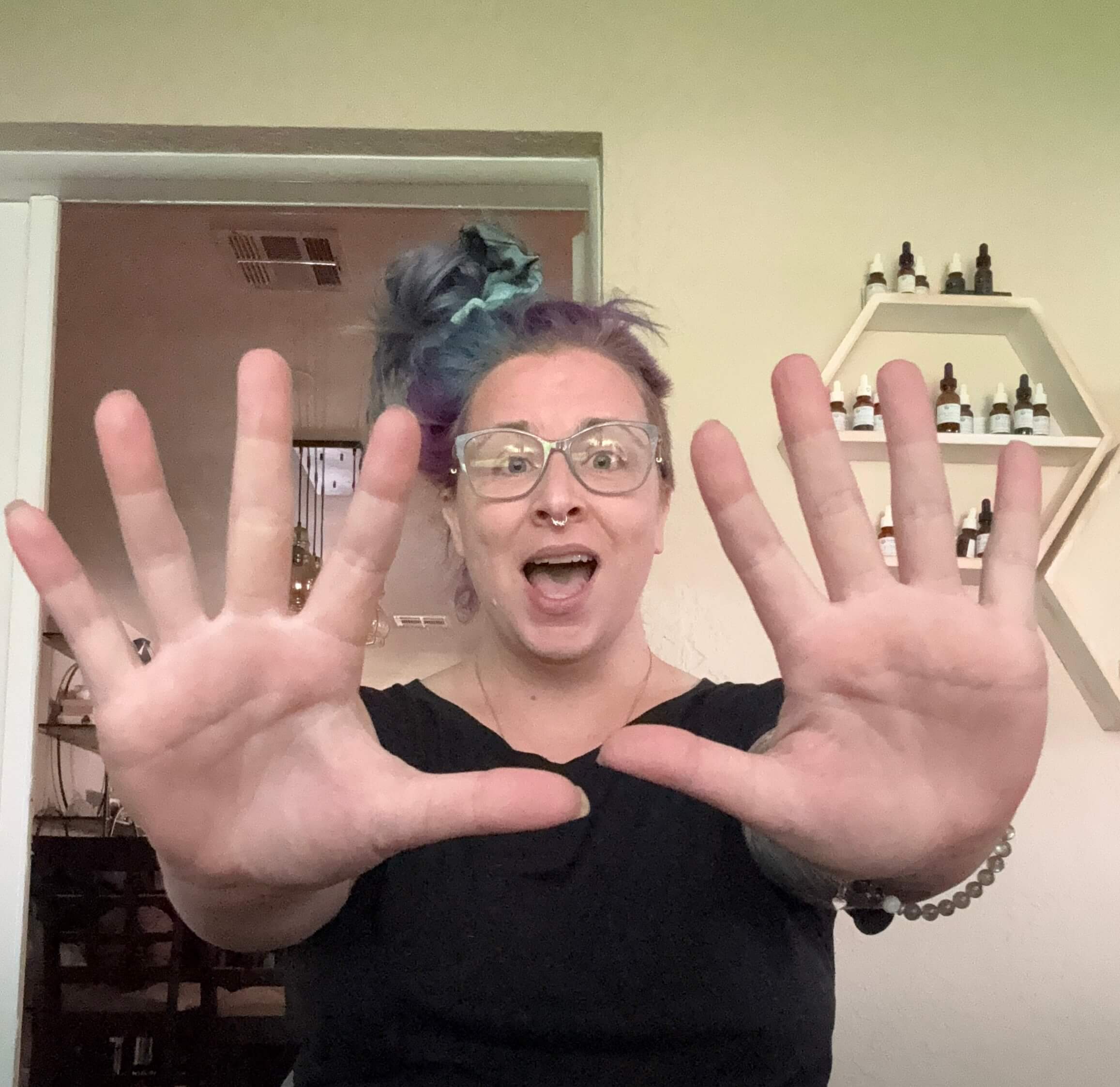 A person with glasses and colorful hair holds both hands up toward the camera, fingers spread wide, and smiles excitedly. Shelves with bottles are visible on the wall behind them.