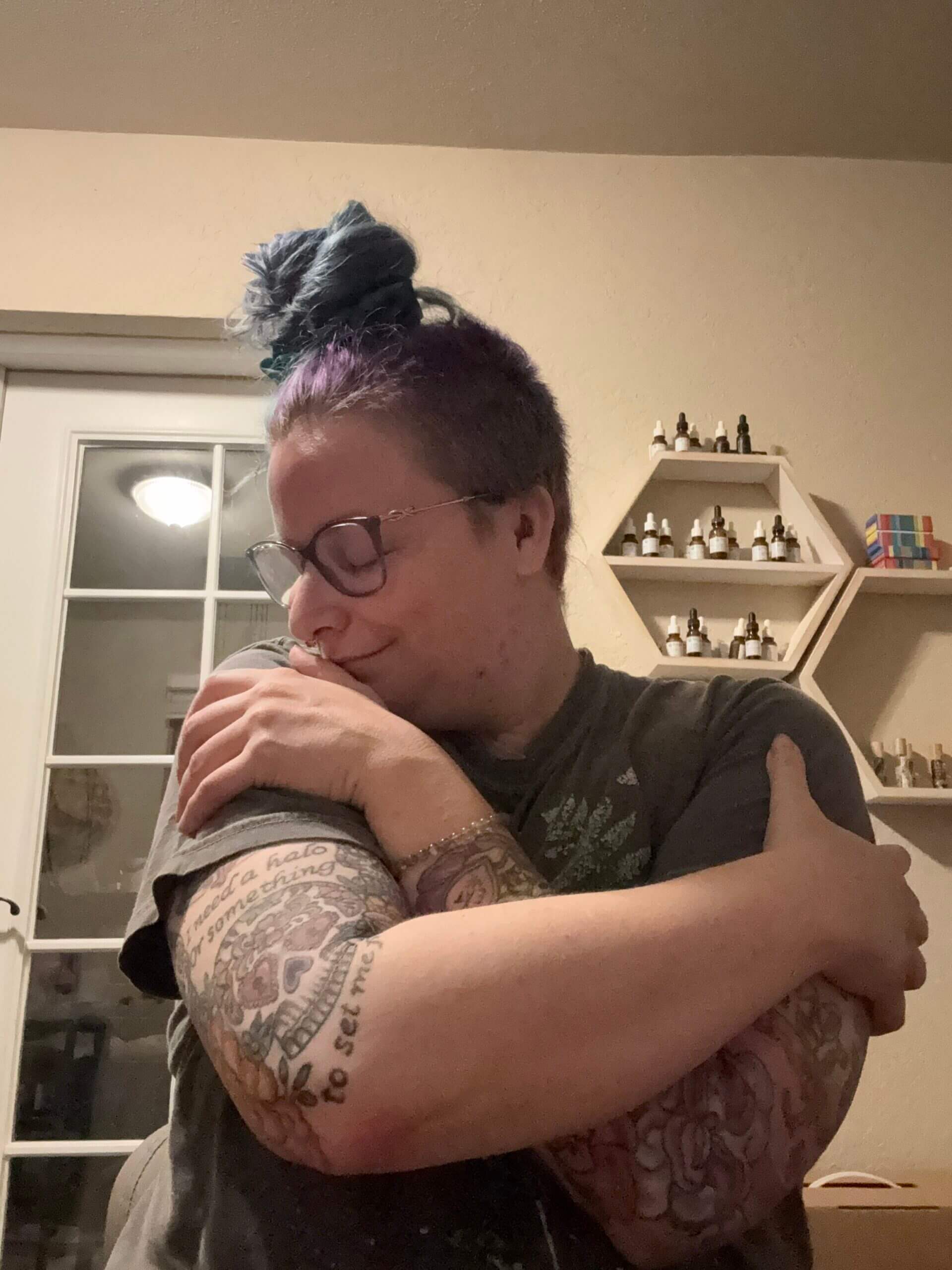 A person with glasses, blue and purple hair in a bun, and tattooed arms hugs themselves while smiling softly, standing in a cozy room with shelves and bottles in the background.