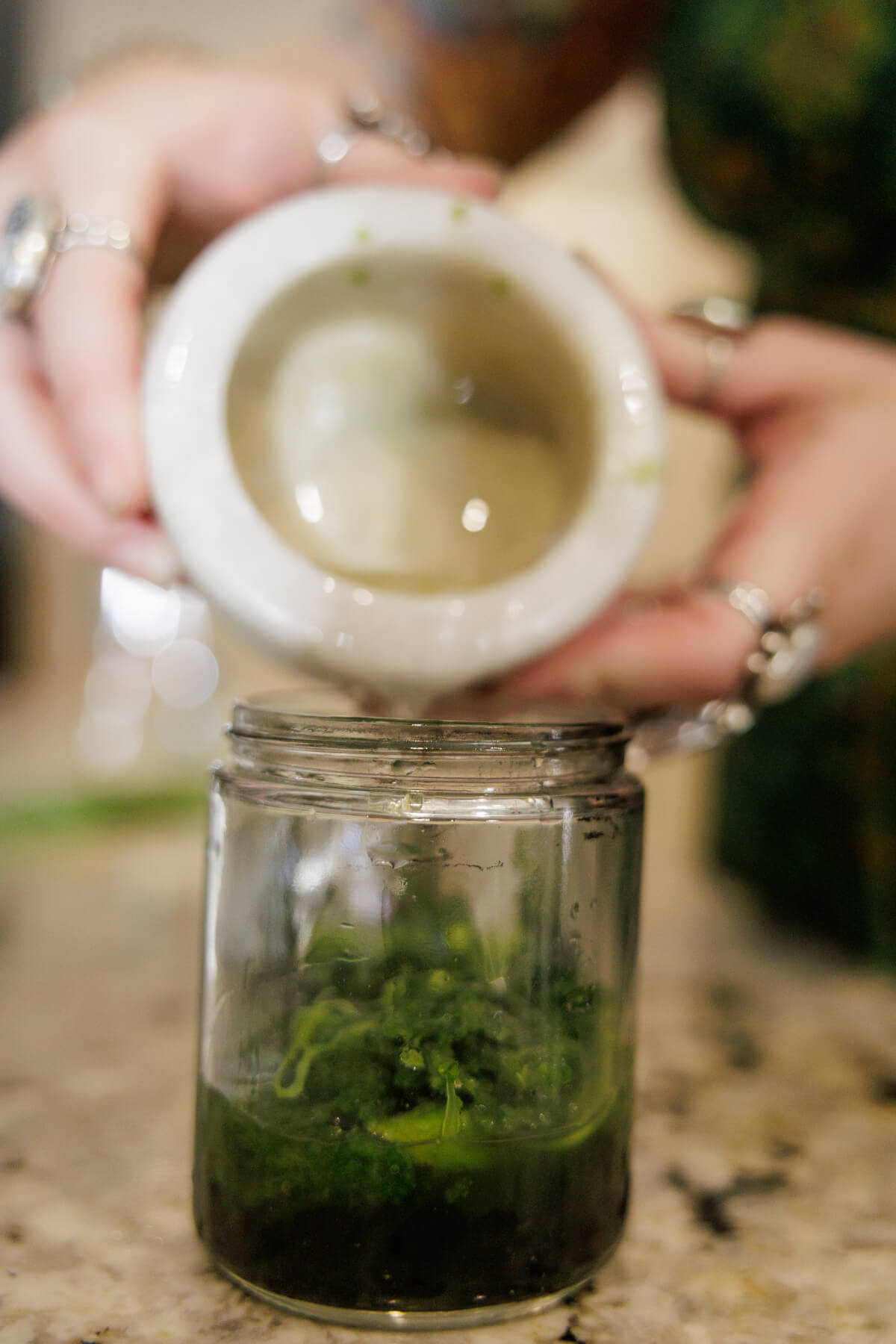 A person holds a mortar above a glass jar containing mashed green herbs or plants, suggesting they are transferring the contents. The scene appears to be on a kitchen counter.