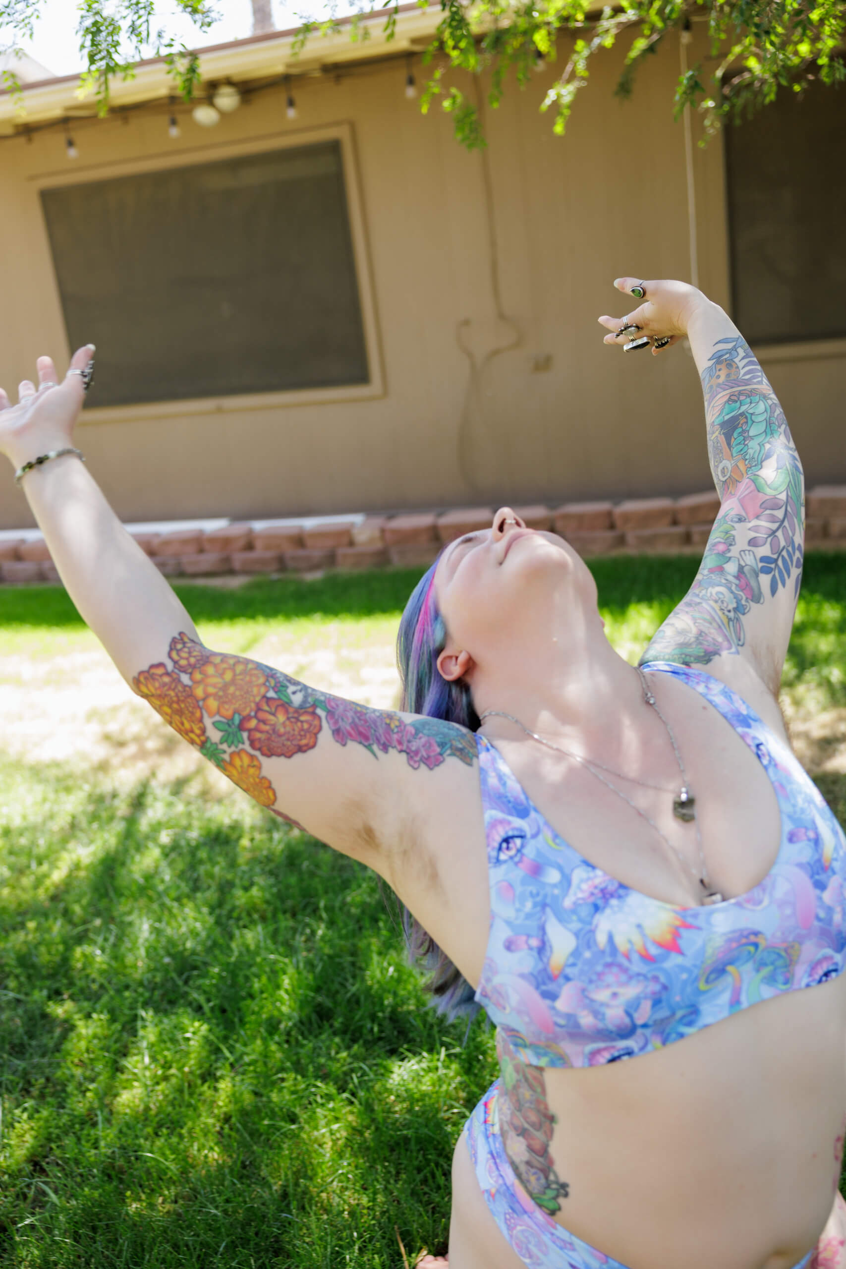 A person in a colorful swimsuit with tattoos on their arms stretches their arms wide and tilts their head back while standing on grass in a sunny yard, with a tan building in the background.