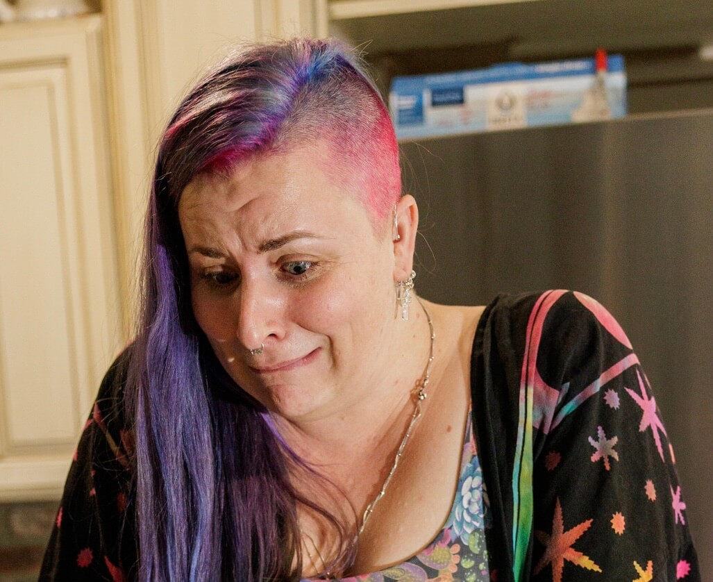 A person with purple and pink hair, wearing a colorful, star-patterned top, looks downward with a worried or uncomfortable expression. The background shows a kitchen with cabinets and a refrigerator.