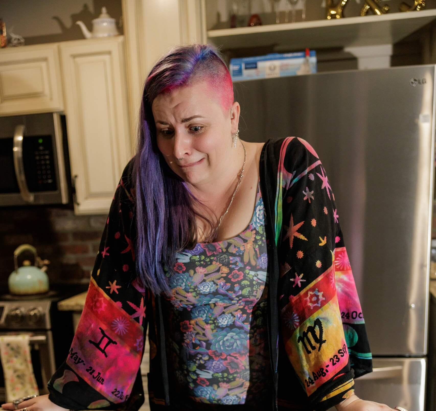 A person with colorful hair and a patterned robe stands in a kitchen, looking worried or anxious. Their robe sleeves display zodiac signs and dates.