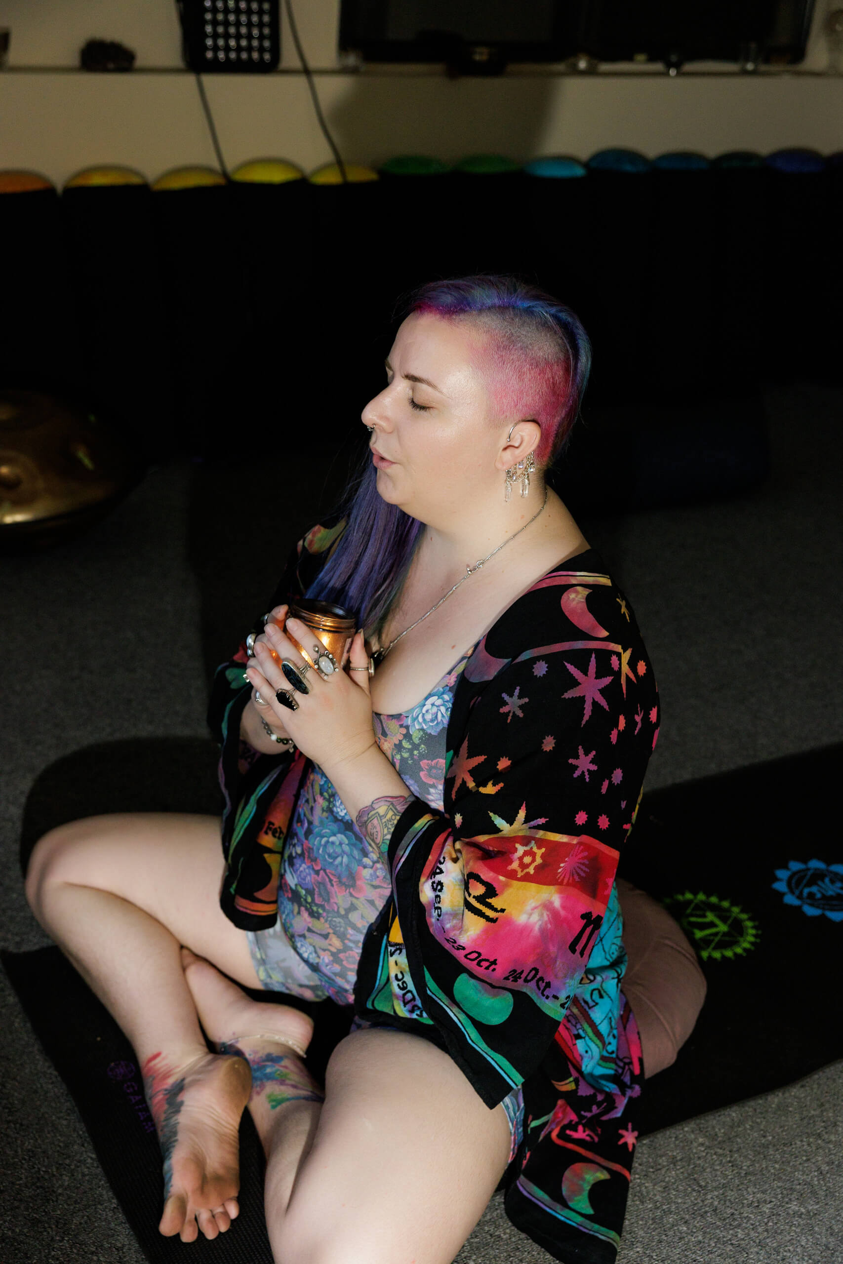 A person with colorful hair and a vibrant robe sits cross-legged on a yoga mat, eyes closed, holding a small cup with both hands in a peaceful, meditative setting.