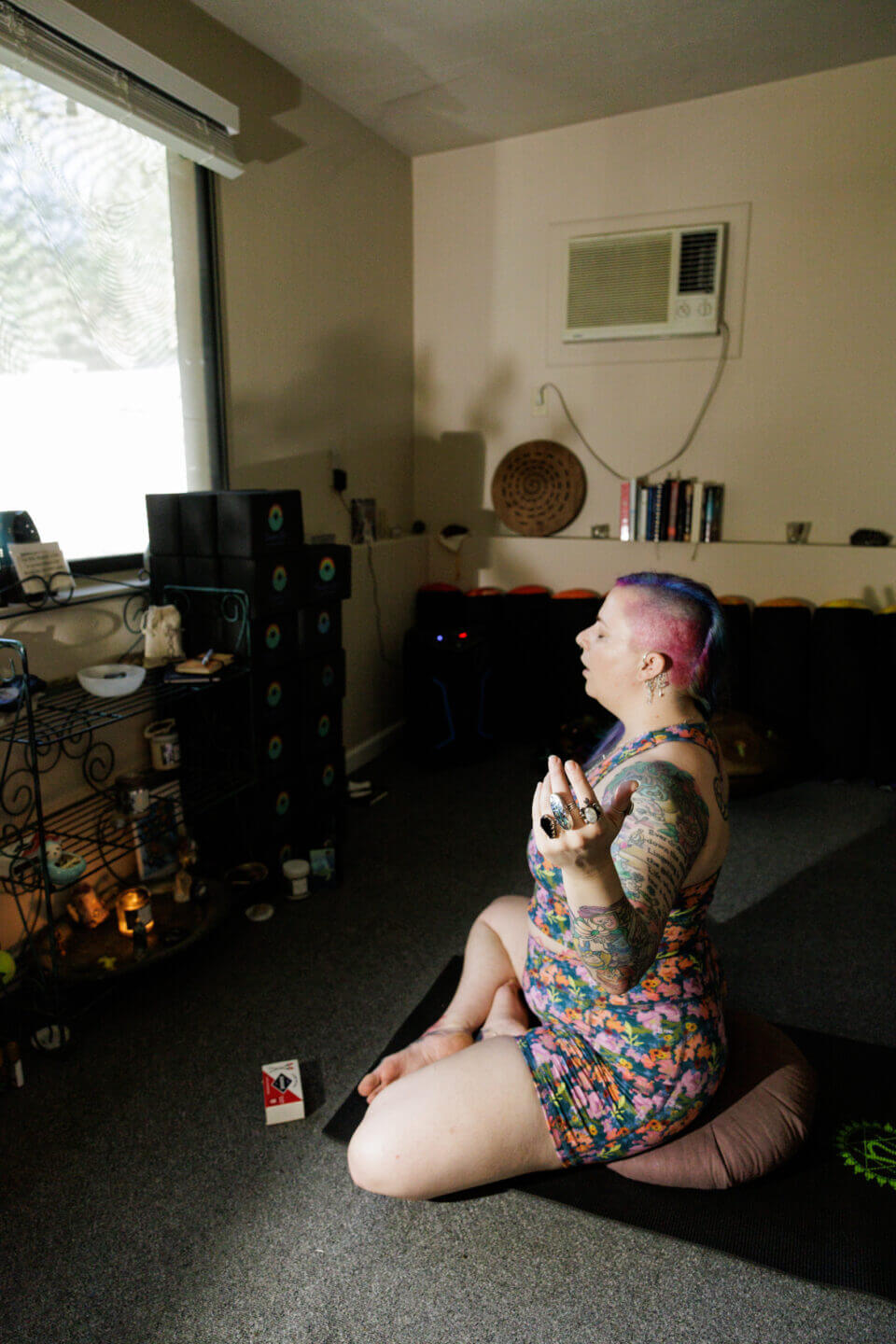 A person with colorful hair and tattoos sits cross-legged on a cushion in a dimly lit room, meditating in front of an altar by a window. The room has shelves with books, candles, and various decorative items.