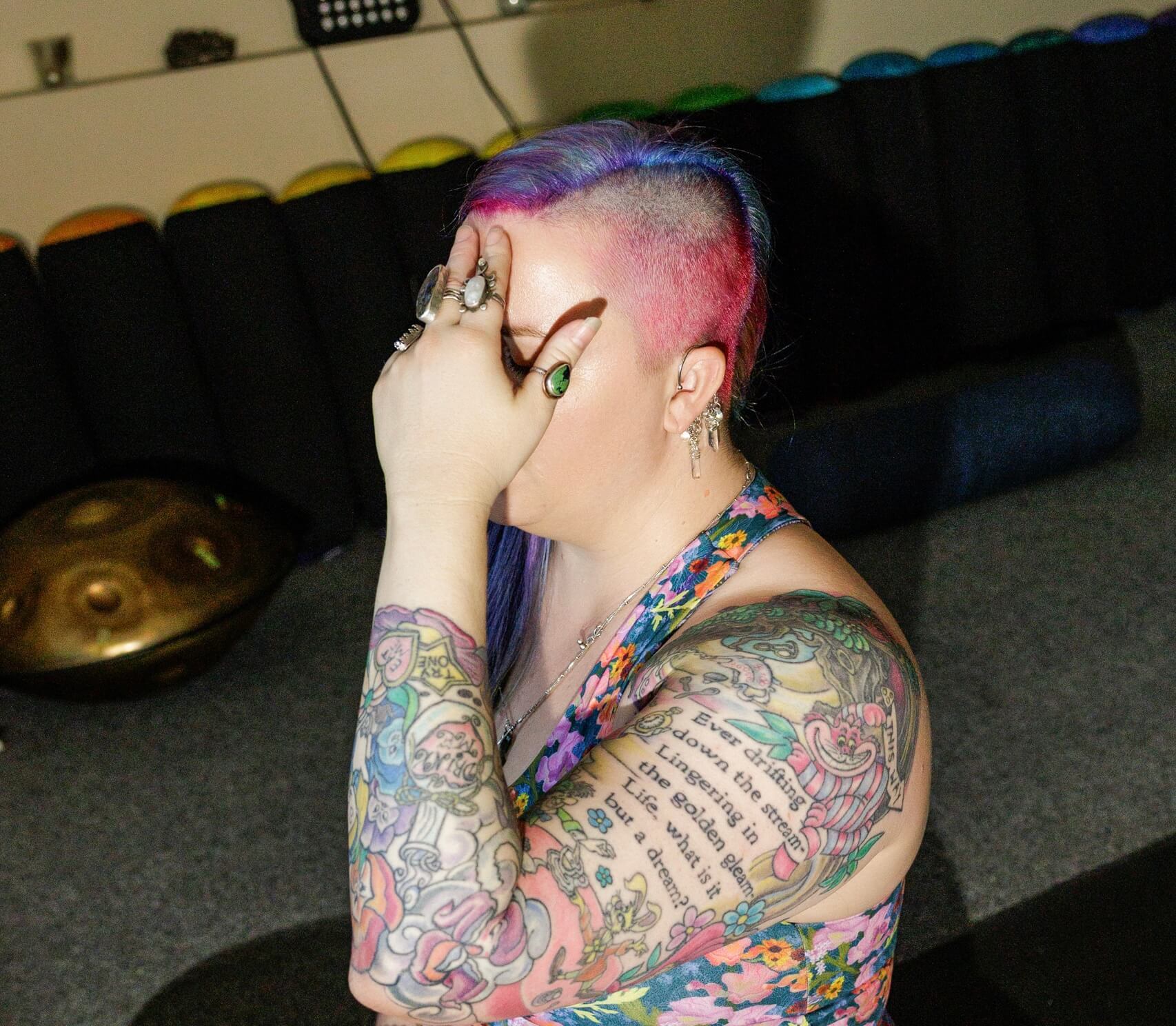 A person with colorful tattoos and half-shaved, pink and purple hair sits indoors, covering their face with one hand. They wear rings and a floral top, and sunlight highlights their tattoos and hair.