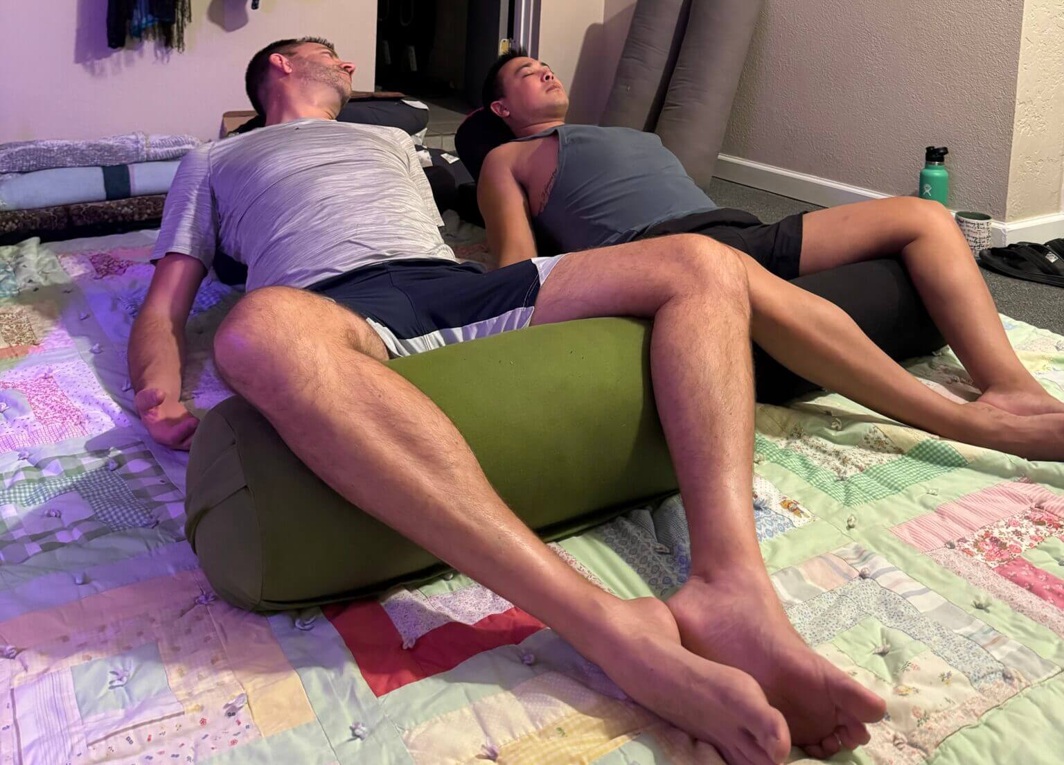 Two people relax indoors on the Deep Love Connection, lying back with cushions supporting their upper bodies and necks, legs stretched out comfortably on the patchwork quilt.