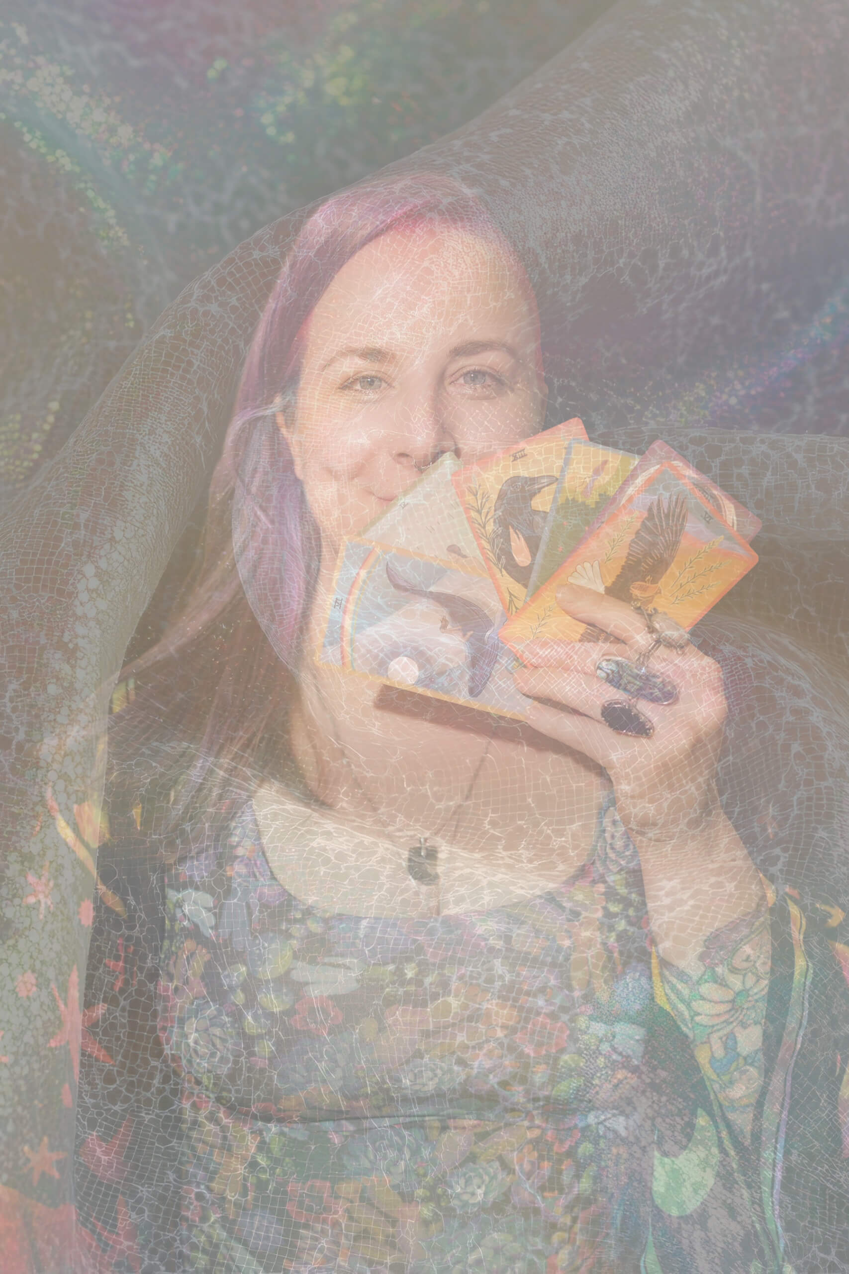 A personwith light-colored hair holds several illustrated tarot cards in front of her face, smiling. She is draped in a sheer, patterned fabric with rainbow hues, creating a mystical and dreamy atmosphere.