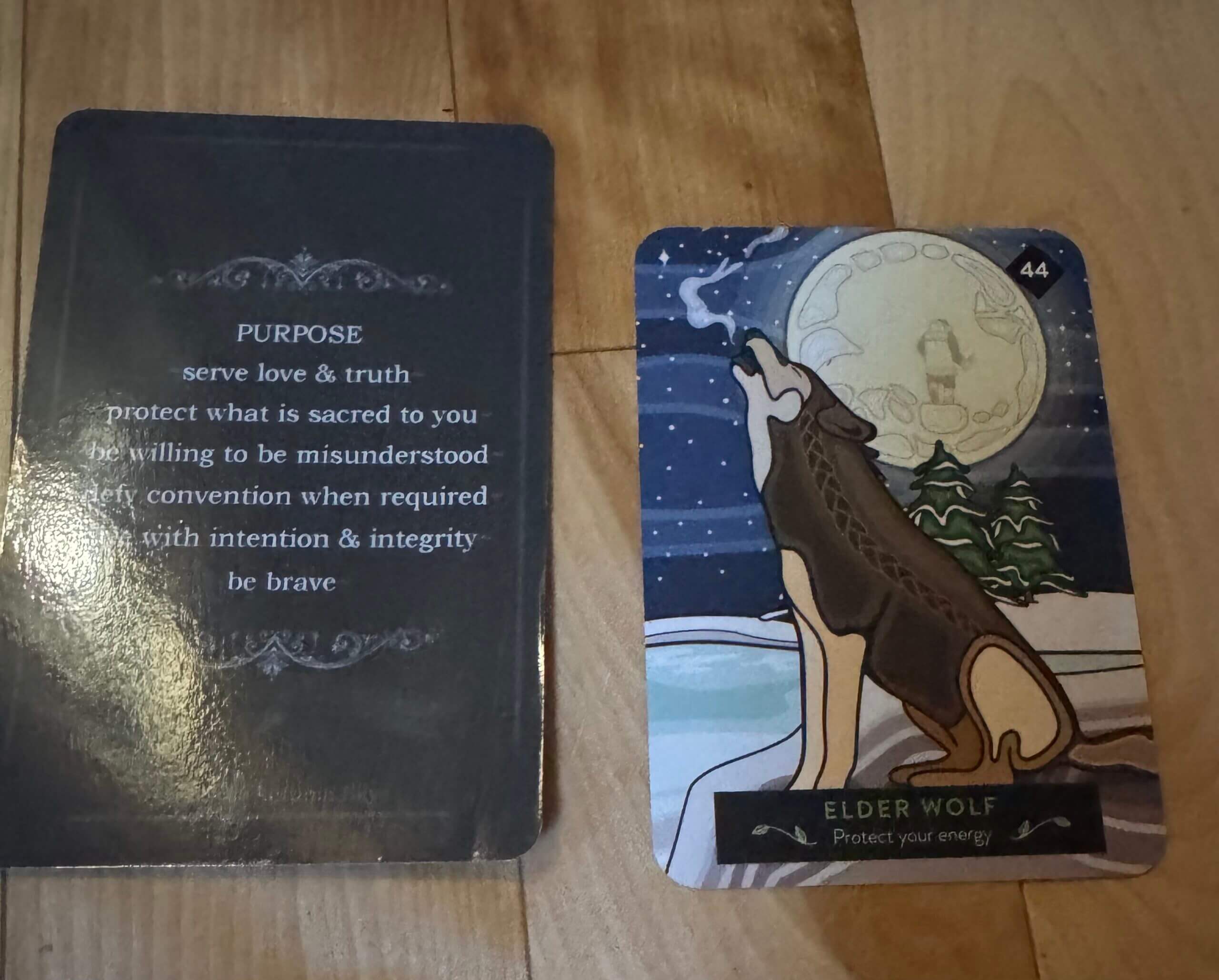 Two cards on a wooden surface. The left card reads about purpose, courage, and integrity. The right card shows a howling wolf under a full moon, with the text Elder Wolf — Protect your energy.