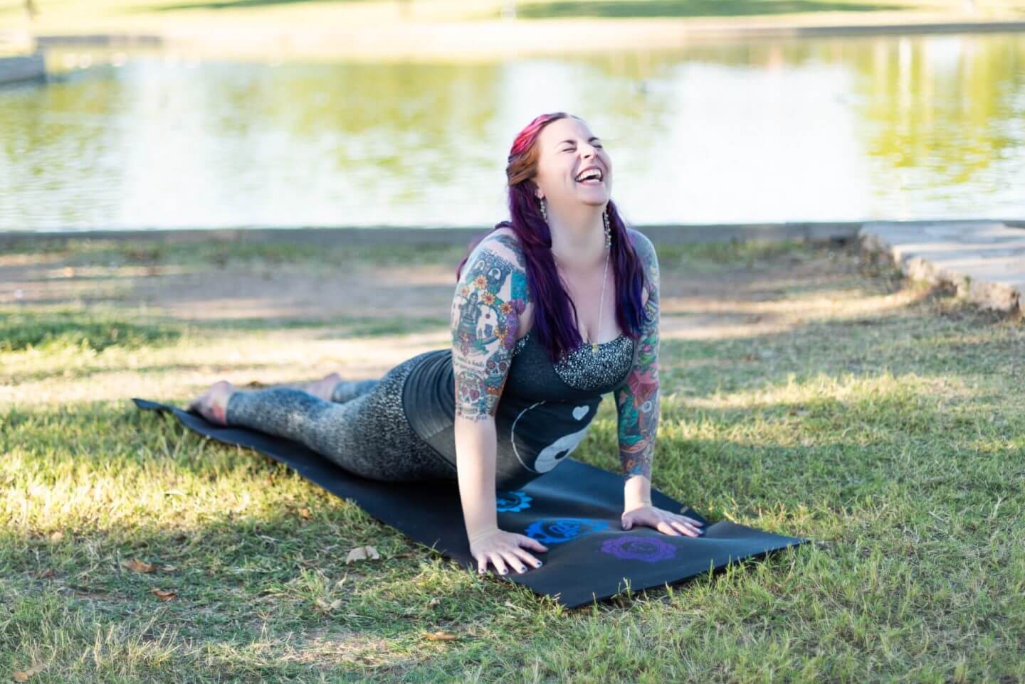 A person with colorful hair and tattoos is practicing yoga outdoors on a grassy area by a lake. They are in the Cobra pose on a black mat, smiling and looking upward. The sun is shining, casting gentle shadows.
