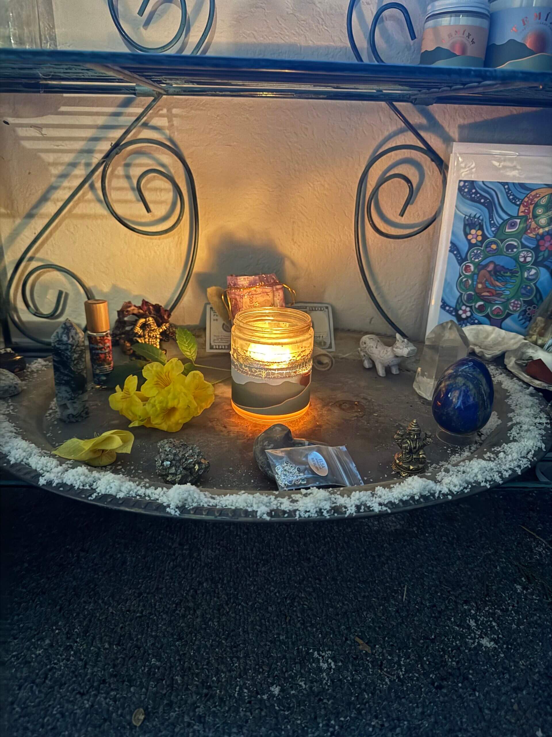 A lit candle sits on a round tray surrounded by crystals, a yellow flower, seashells, a small figurine, and other spiritual items, all placed on a lower shelf with decorative metalwork.