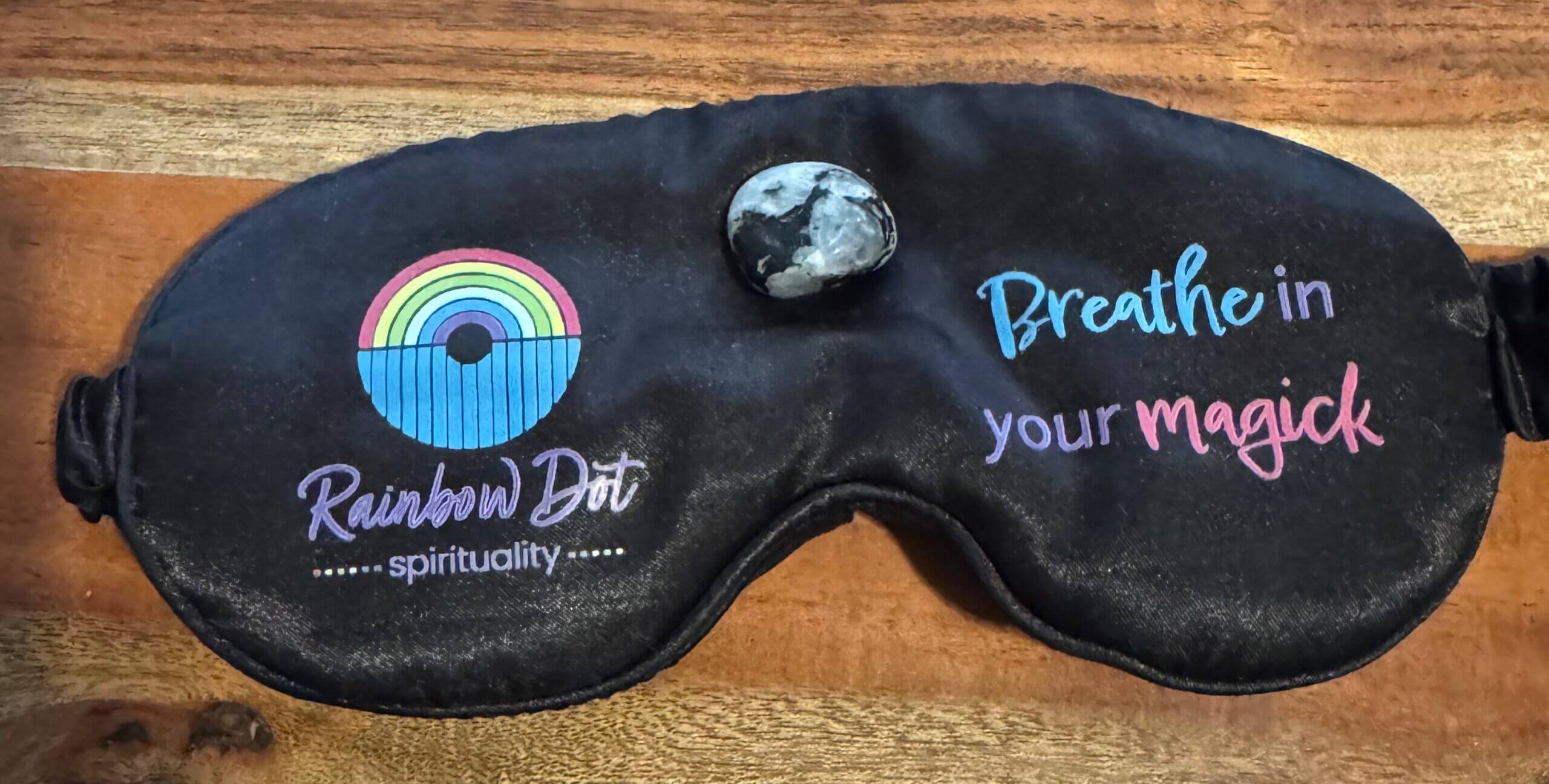 A black sleep mask with a rainbow and dot logo, the words Rainbow Dot spirituality, and the phrase Breathe in your magick in blue and pink, lying on a wooden surface with a small stone on top.