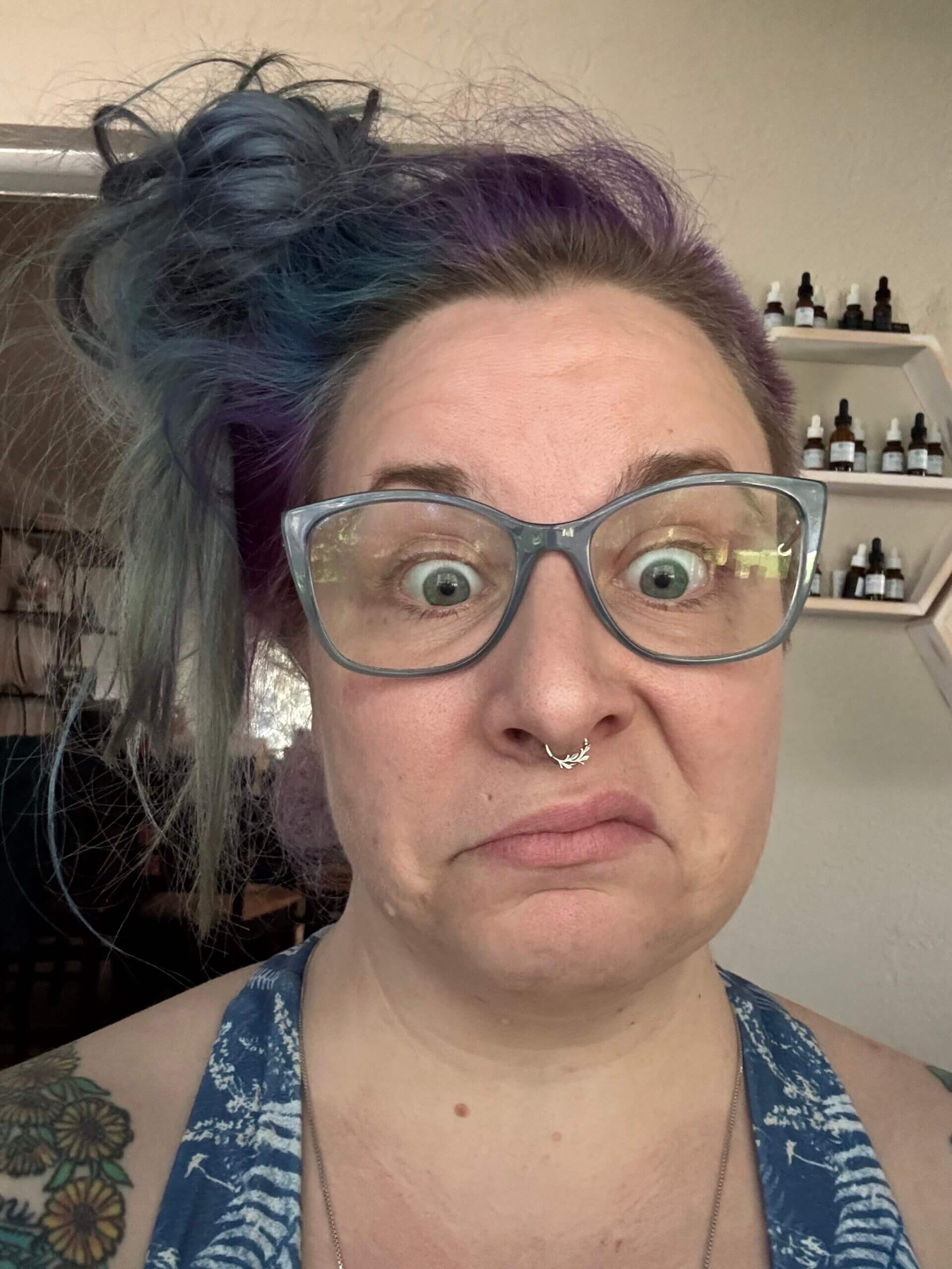 A person with light skin, blue glasses, and purple hair in a messy bun makes a surprised, skeptical face. They wear a blue patterned top and have a nose ring. Shelves with bottles are visible in the background.