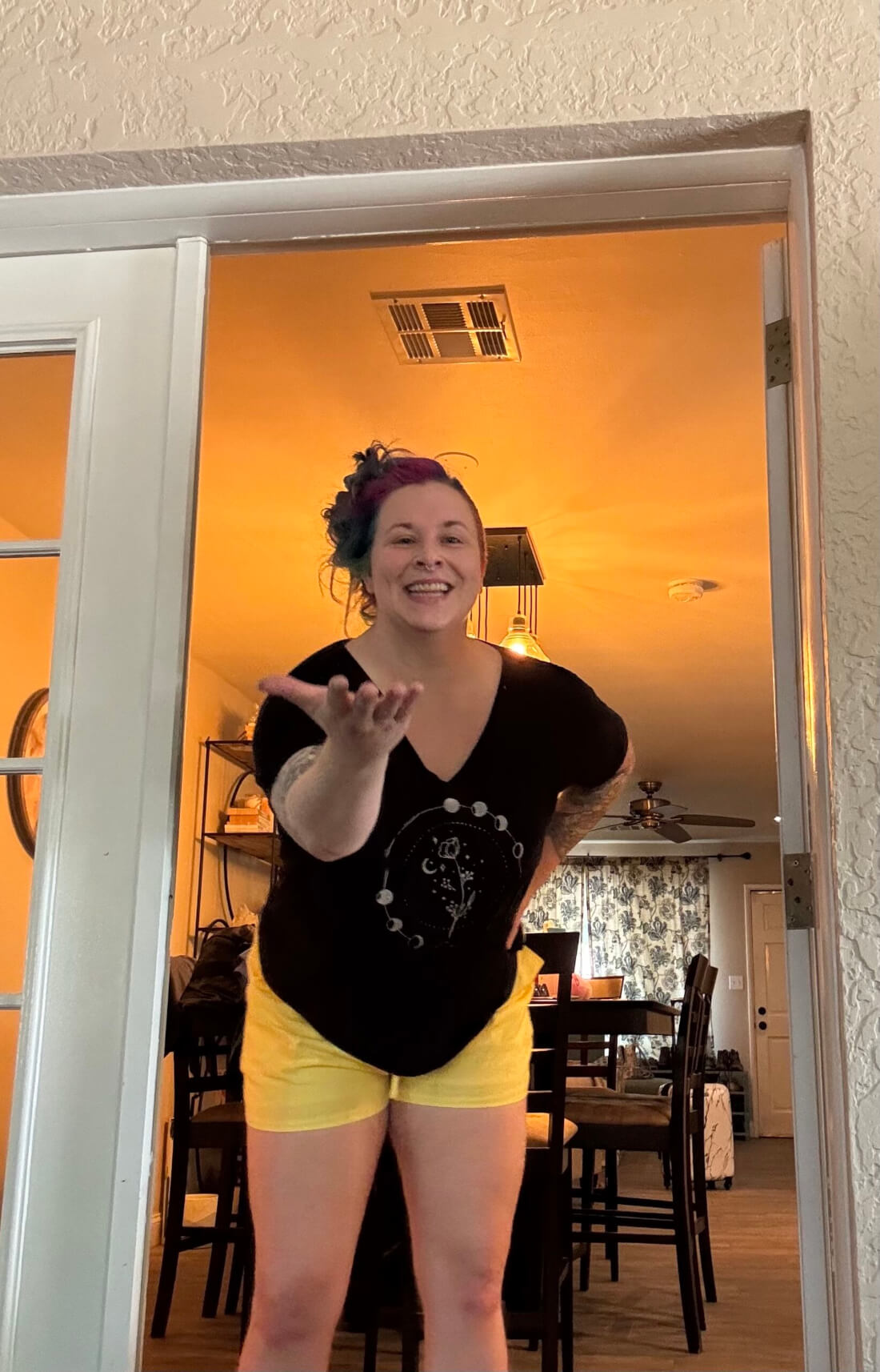 A person with colorful hair, wearing a black shirt and yellow shorts, stands in a doorway and blows a kiss toward the camera. The room behind them is warmly lit, with a dining table and ceiling fan visible.