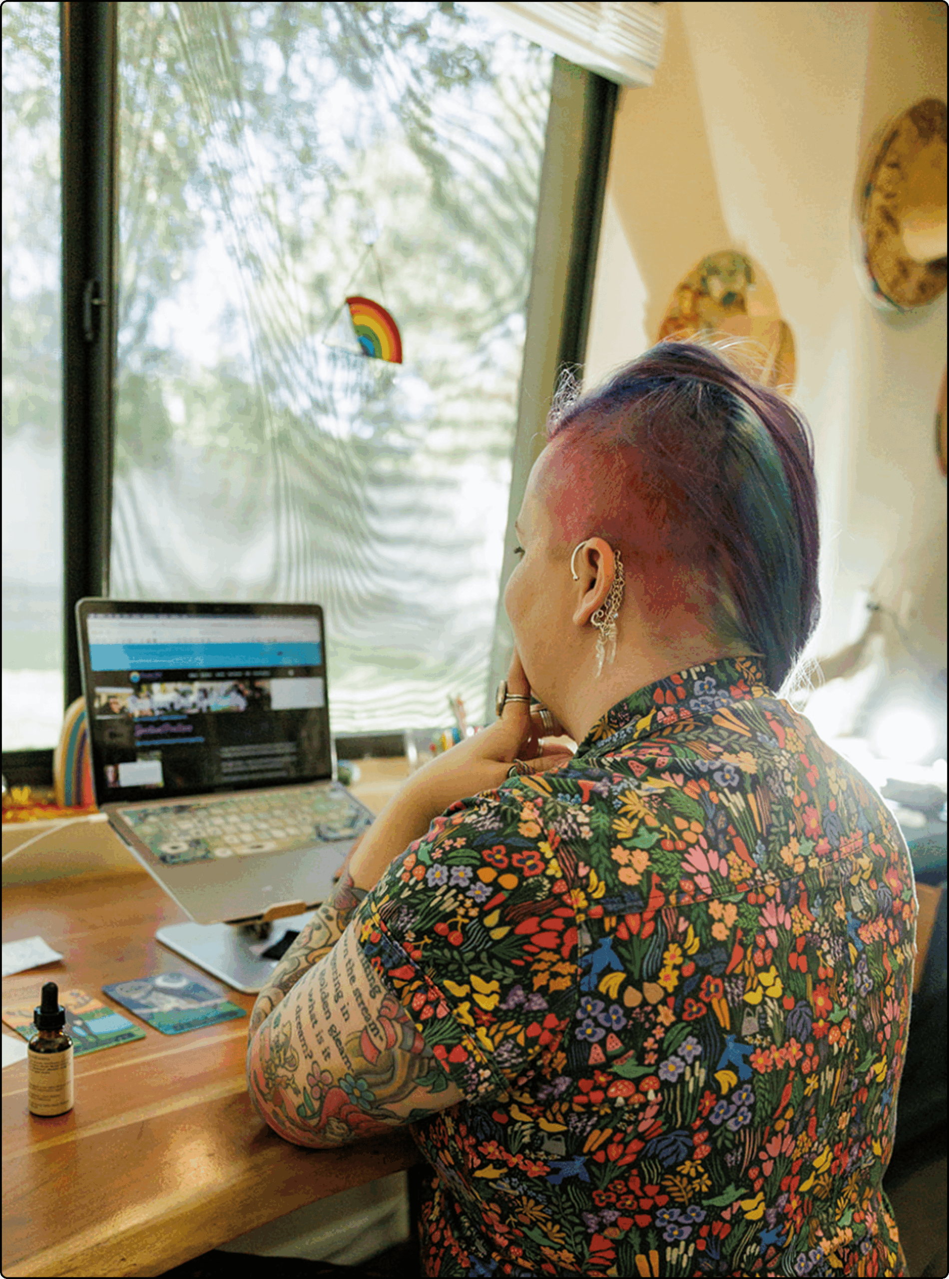 A person with colorful hair and tattoos sits at a desk, working on a laptop. They wear a floral shirt and are facing a window with a rainbow decoration. Light streams in, and various objects are on the wooden desk.