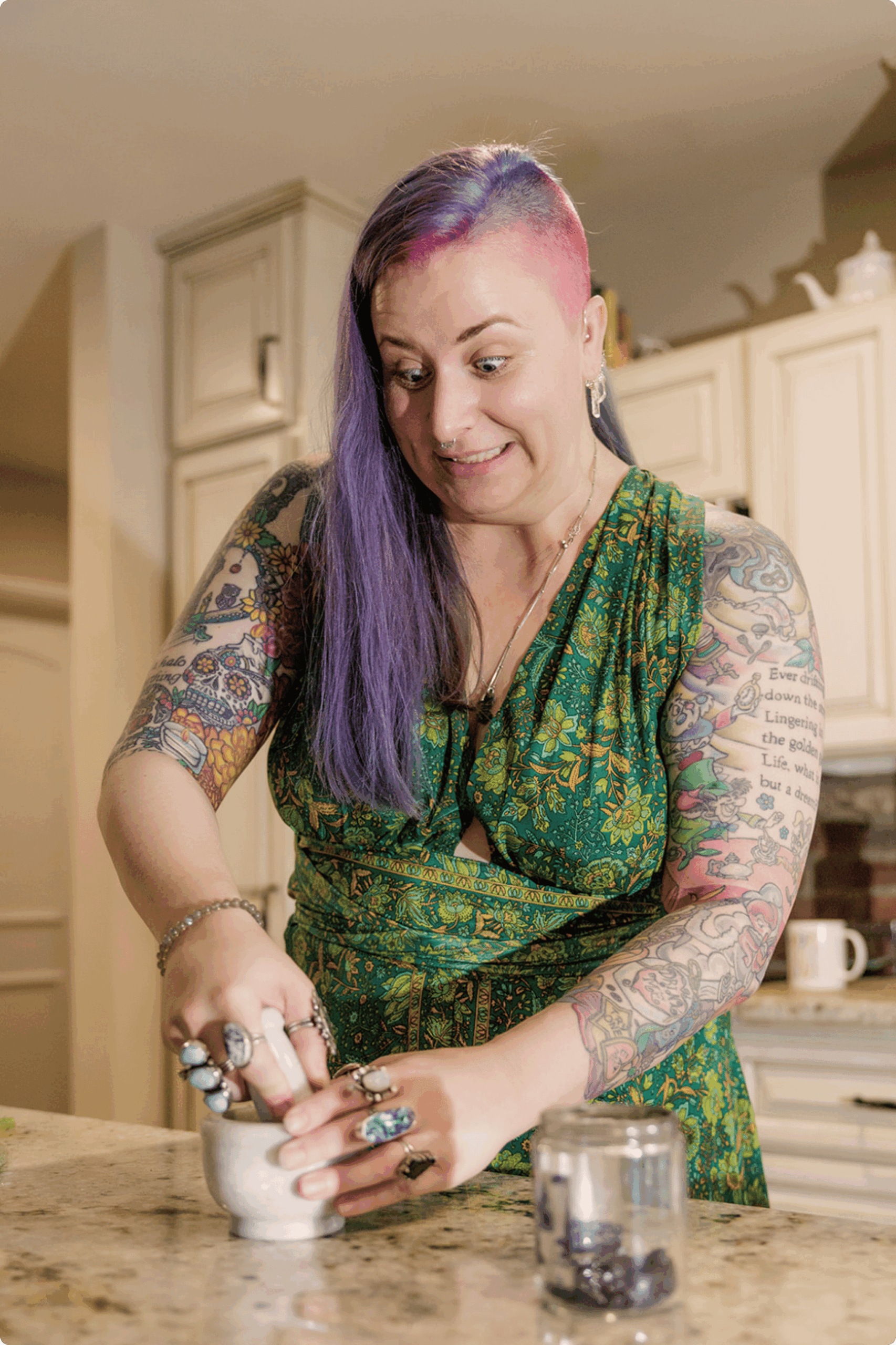 A person with purple hair, tattoos, and rings is smiling while using a mortar and pestle to crush something on a kitchen counter. A glass jar with dark berries sits nearby.