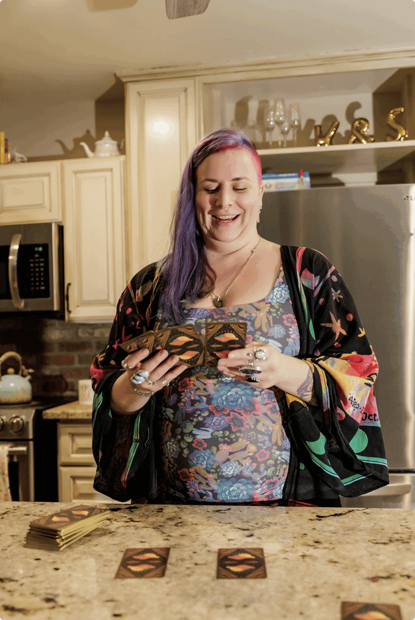 A person with long purple hair stands in a kitchen, smiling while shuffling tarot cards. They wear a floral dress and a colorful robe, with rings on their fingers. Tarot cards are spread out on the countertop.