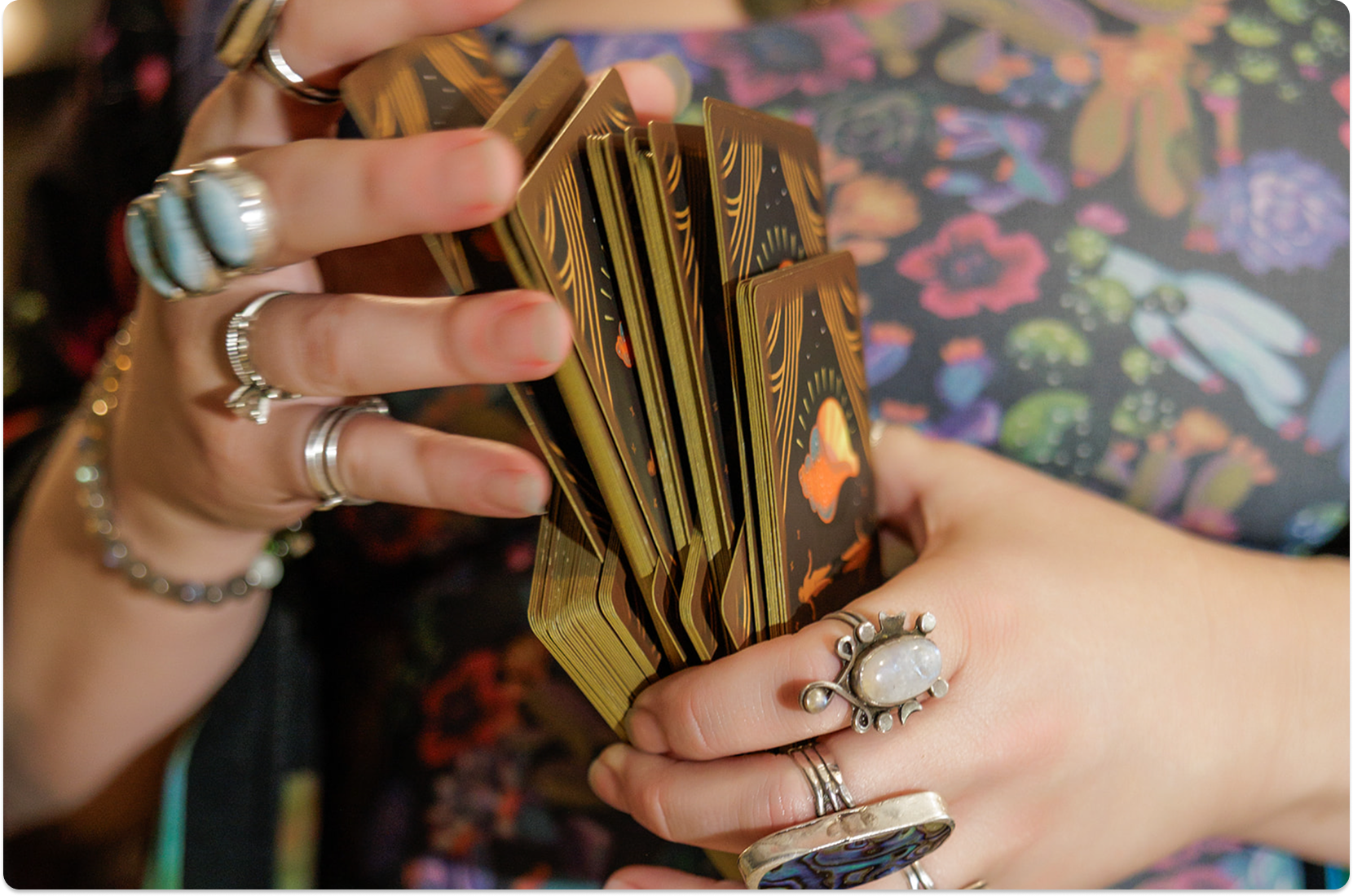 A person wearing multiple rings shuffles a deck of tarot cards, with colorful, floral-patterned clothing visible in the background.