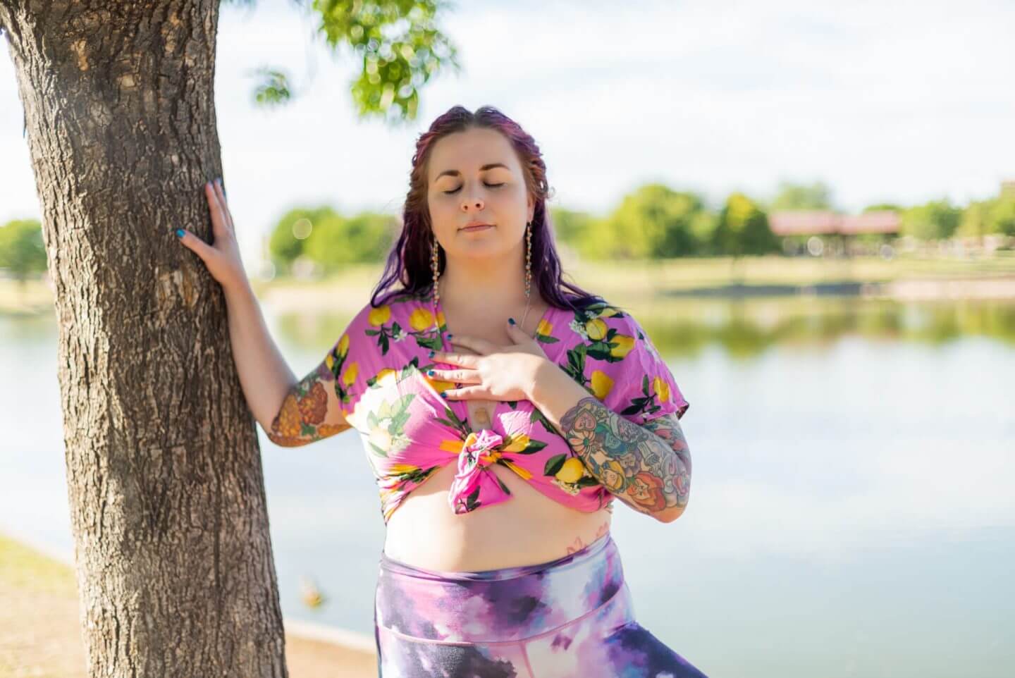 A person with colorful tattoos stands outdoors by a lake, wearing a floral top and purple leggings. They have one hand on a tree and the other on their chest, with eyes closed, appearing relaxed and serene.