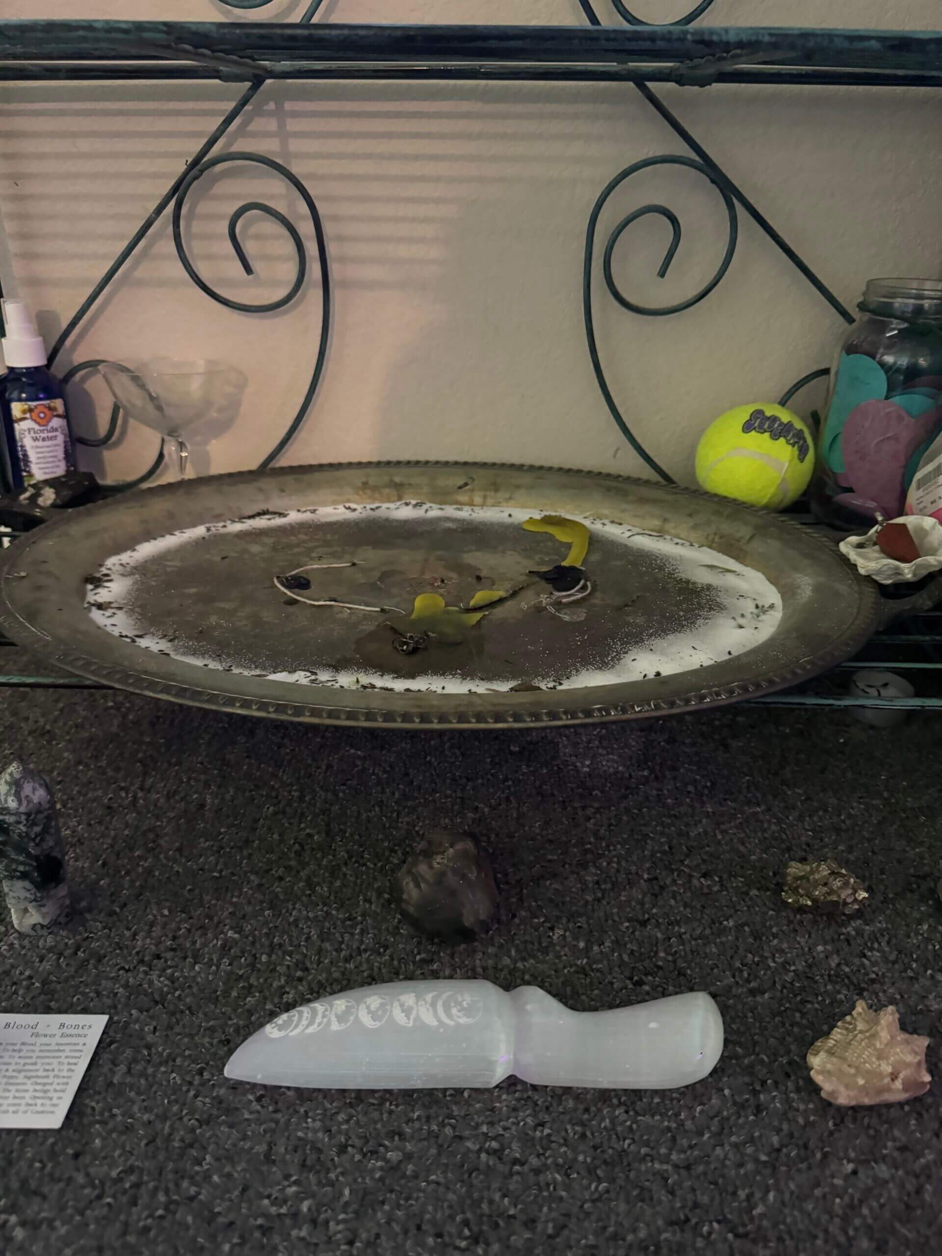 A metal tray with a candle, surrounded by salt and leaves, sits on a shelf. Nearby are crystals, a white selenite knife, a tennis ball, and a jar filled with colorful objects. The setting has mystical and decorative items.