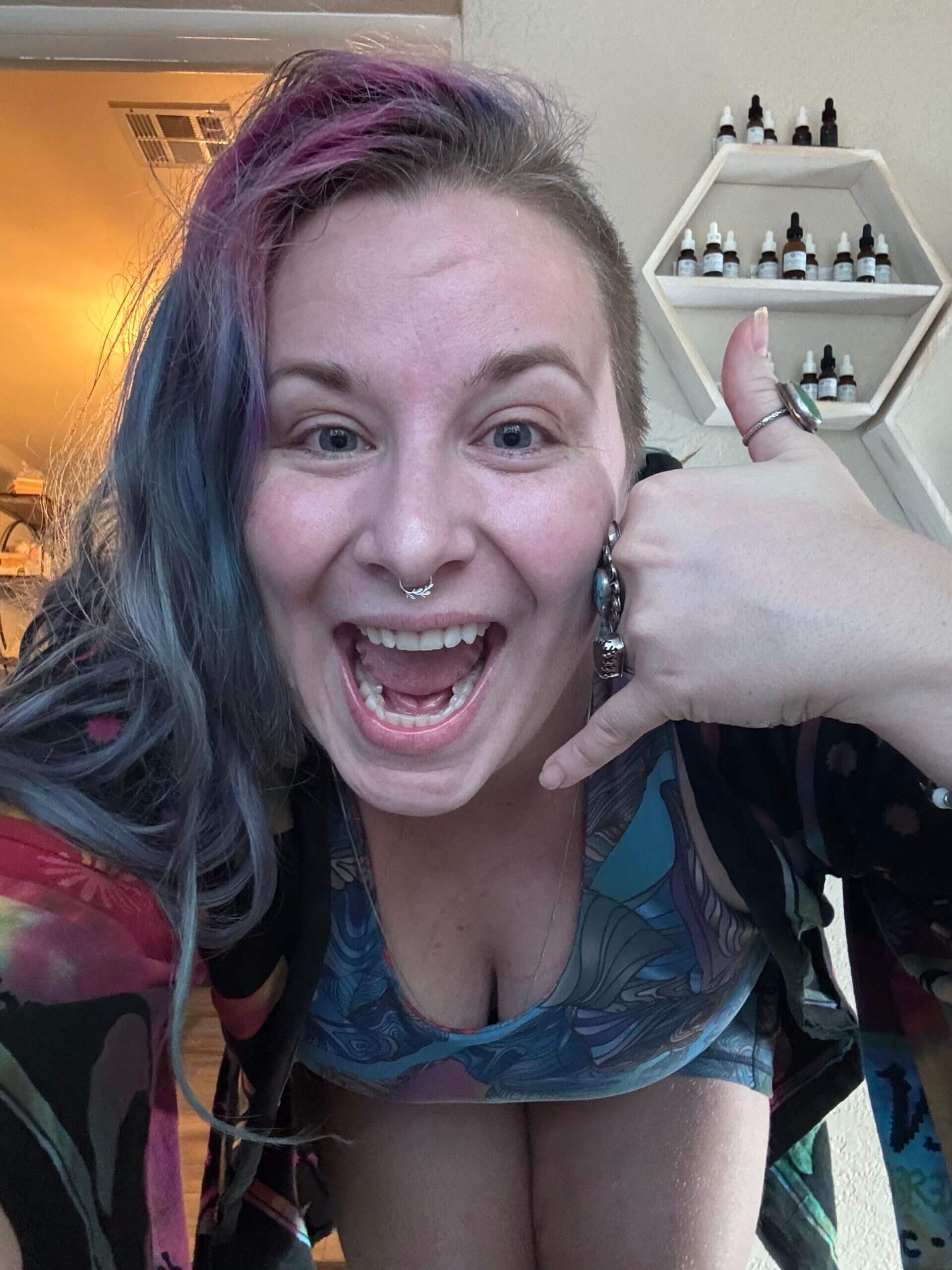 A person with colorful hair and a nose ring smiles widely, making a call me hand gesture. They wear a patterned top and stand indoors near a wall shelf with small bottles.