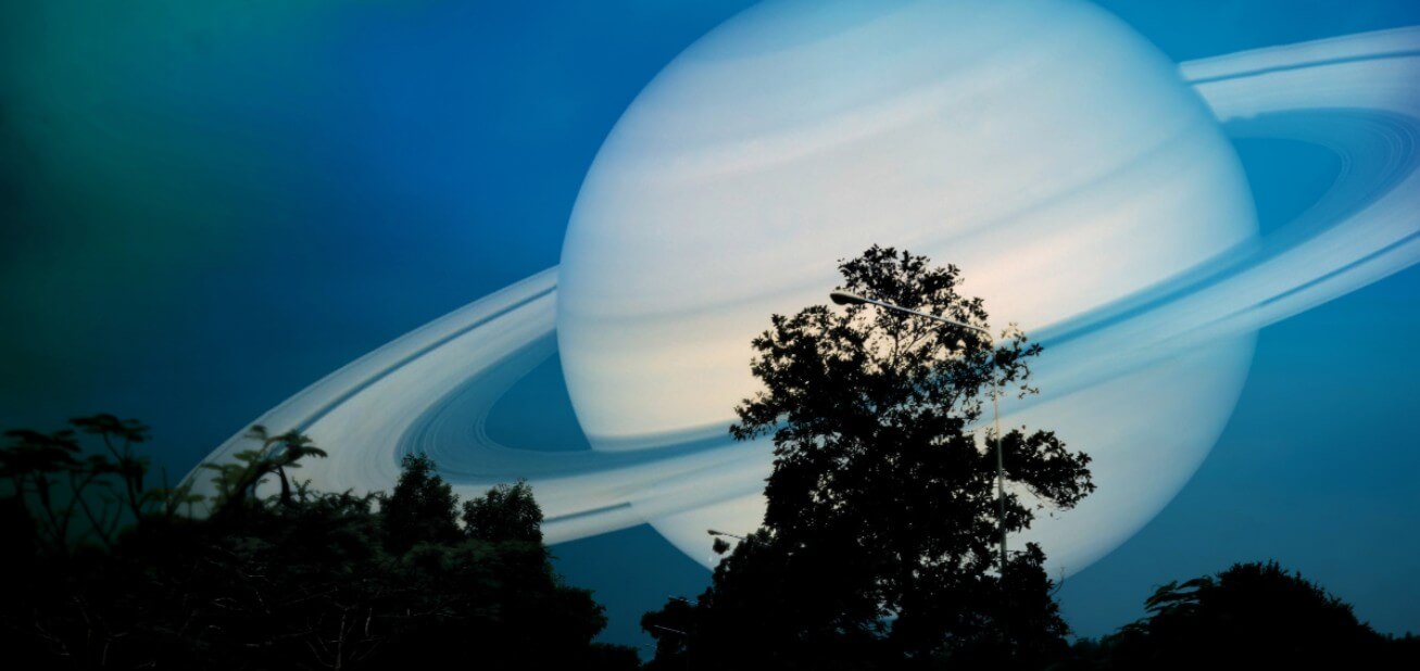 A large tree silhouette stands against a blue sky with an enormous, detailed planet Saturn and its rings looming in the background.