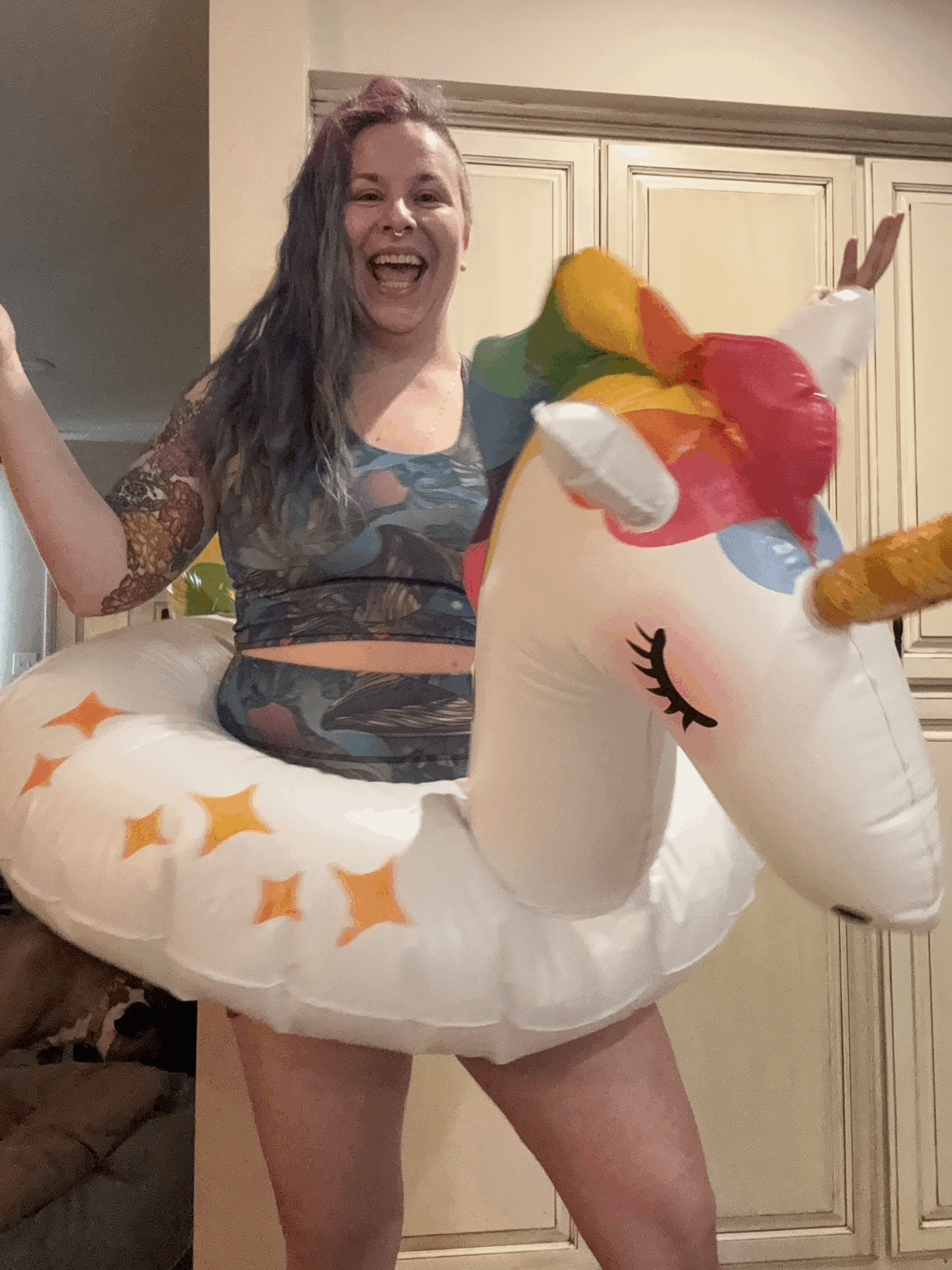 A smiling person with long, wavy hair stands indoors wearing a crop top and shorts, with a large inflatable unicorn pool float around their waist. Their arms are outstretched in a playful pose.