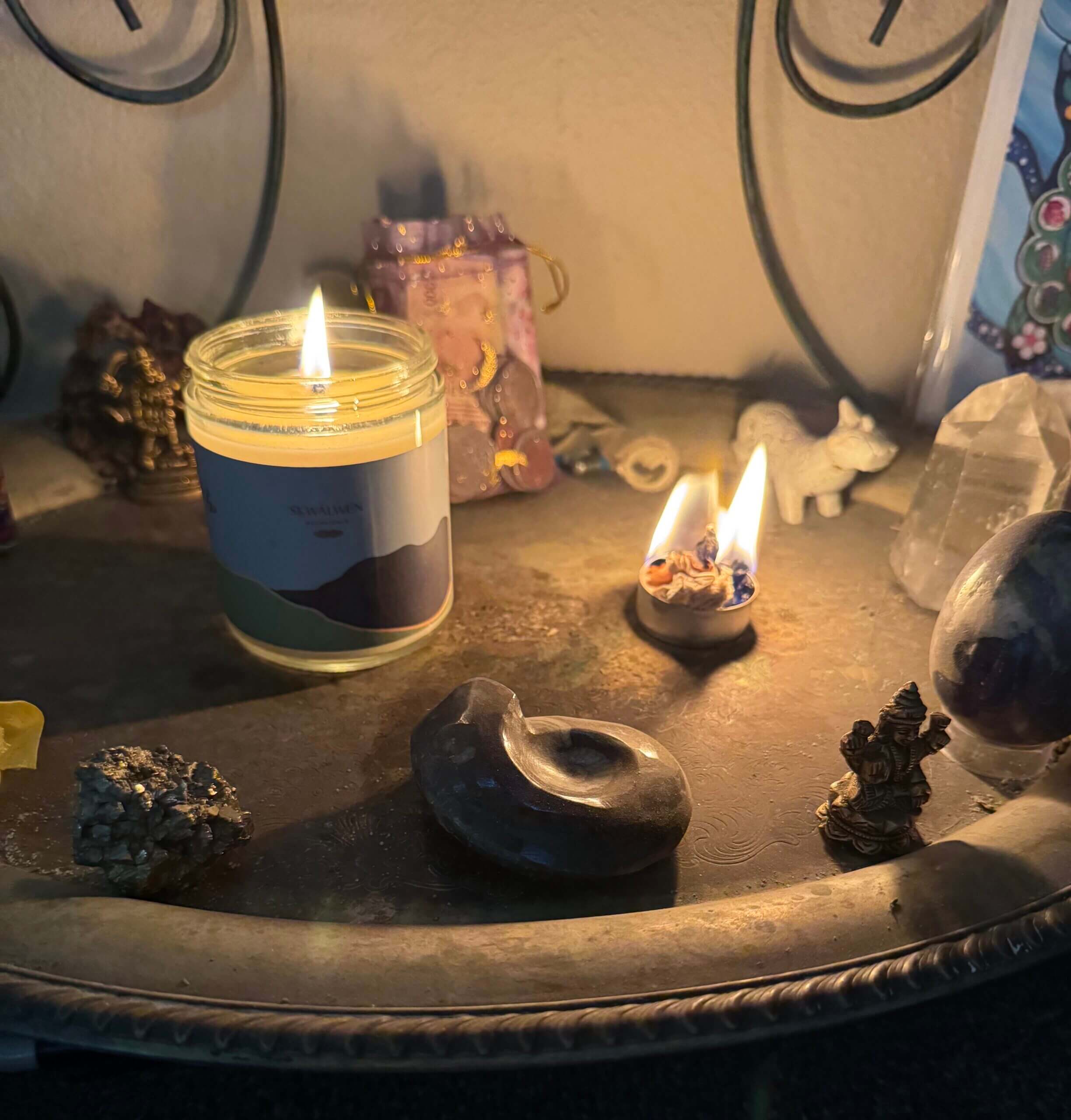 A small altar features a lit candle, two small oil lamps with flames, crystals, a stone sculpture, a decorative bag, and tiny figurines, all arranged on a metal tray against a wall.