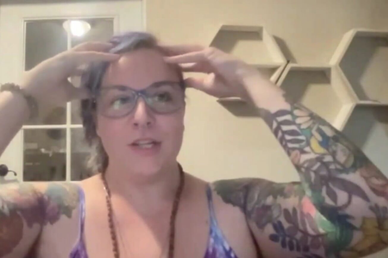 A person with glasses and colorful, tattooed arms sits indoors, touching their forehead with both hands. Hexagonal shelves are visible on the wall behind them.